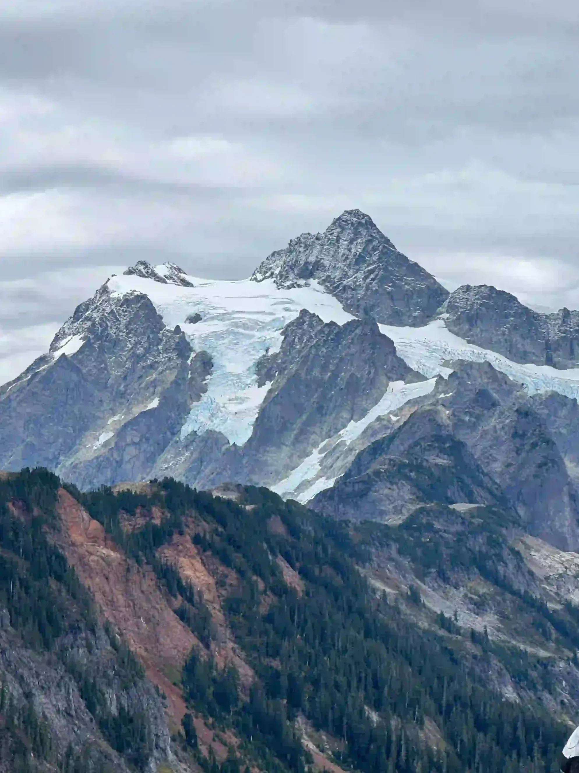 Snow-capped mountain peaks in Seattle, WA with rugged rock formations and dense forested slopes beneath cloudy skies.
