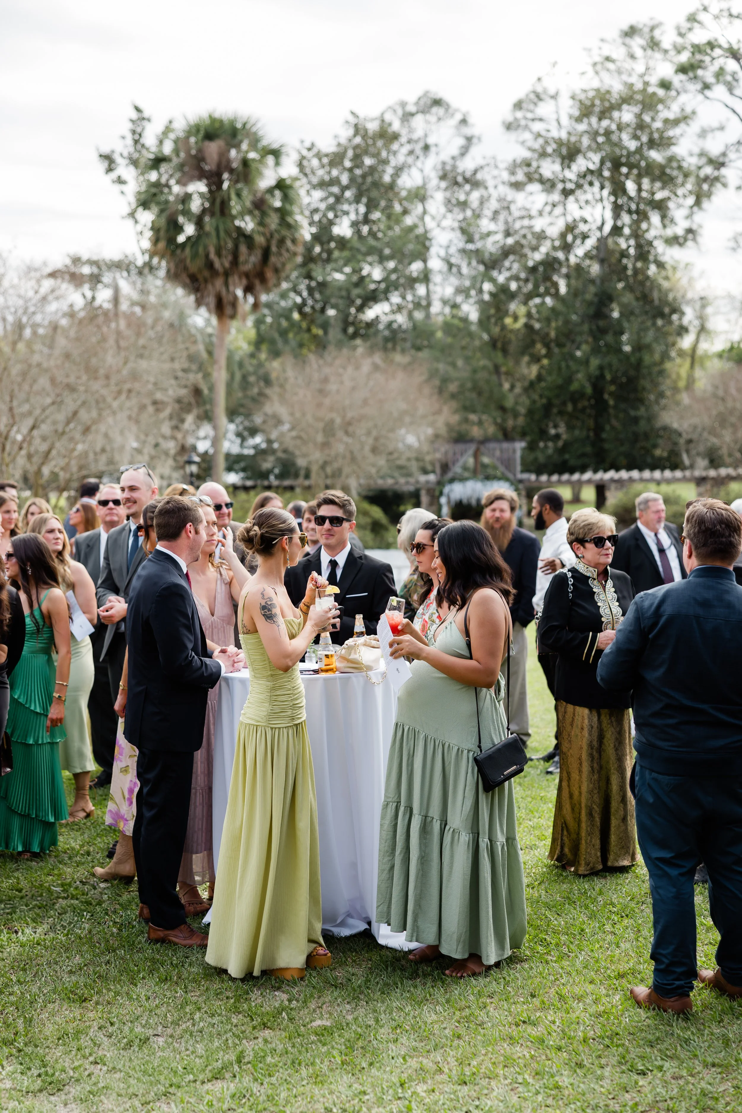 People gathered at an outdoor party or wedding reception with drinks, socializing around a tall cocktail table on a grassy area with trees and a cloudy sky in the background.