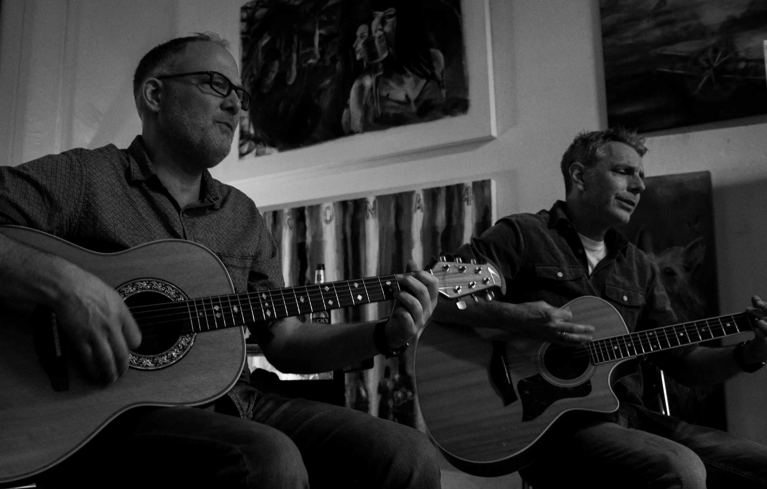 Two men playing acoustic guitars and singing, sitting indoors with artwork hanging on the wall behind them.