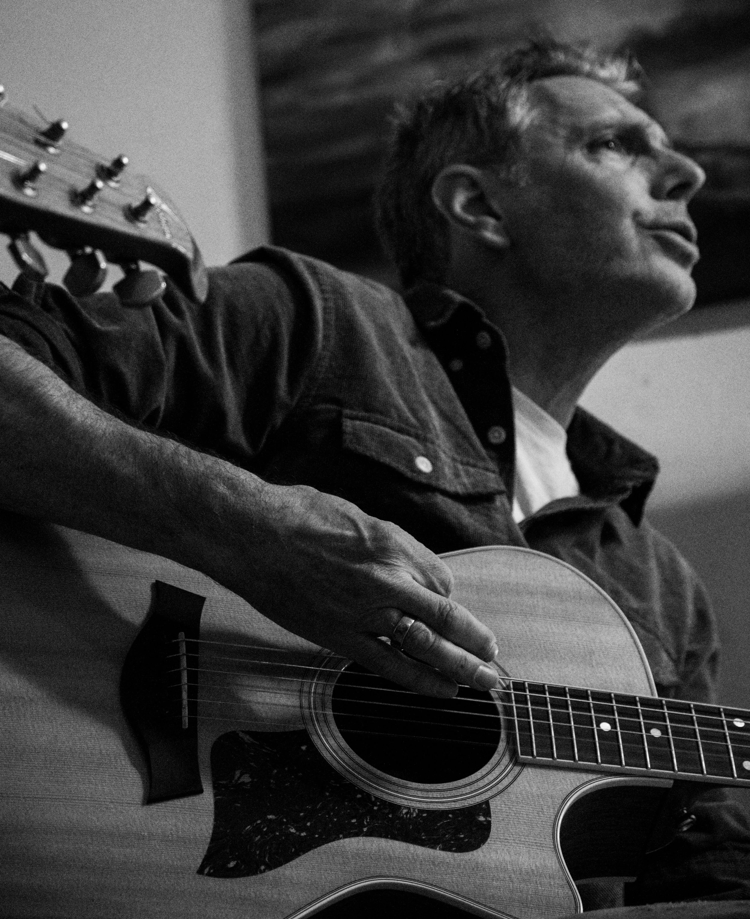 A person playing an acoustic guitar with a close-up of their face and hand on the guitar strings, in black and white.