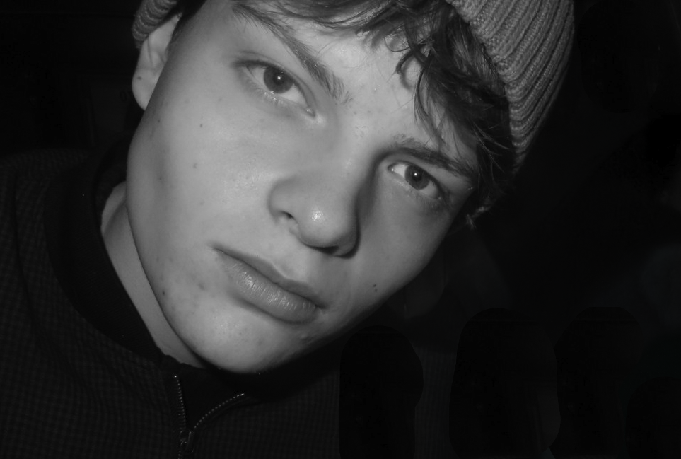 Close-up of a young man with light skin, freckles, and light-colored eyes, wearing a beanie and a dark jacket, looking directly at the camera in black and white.