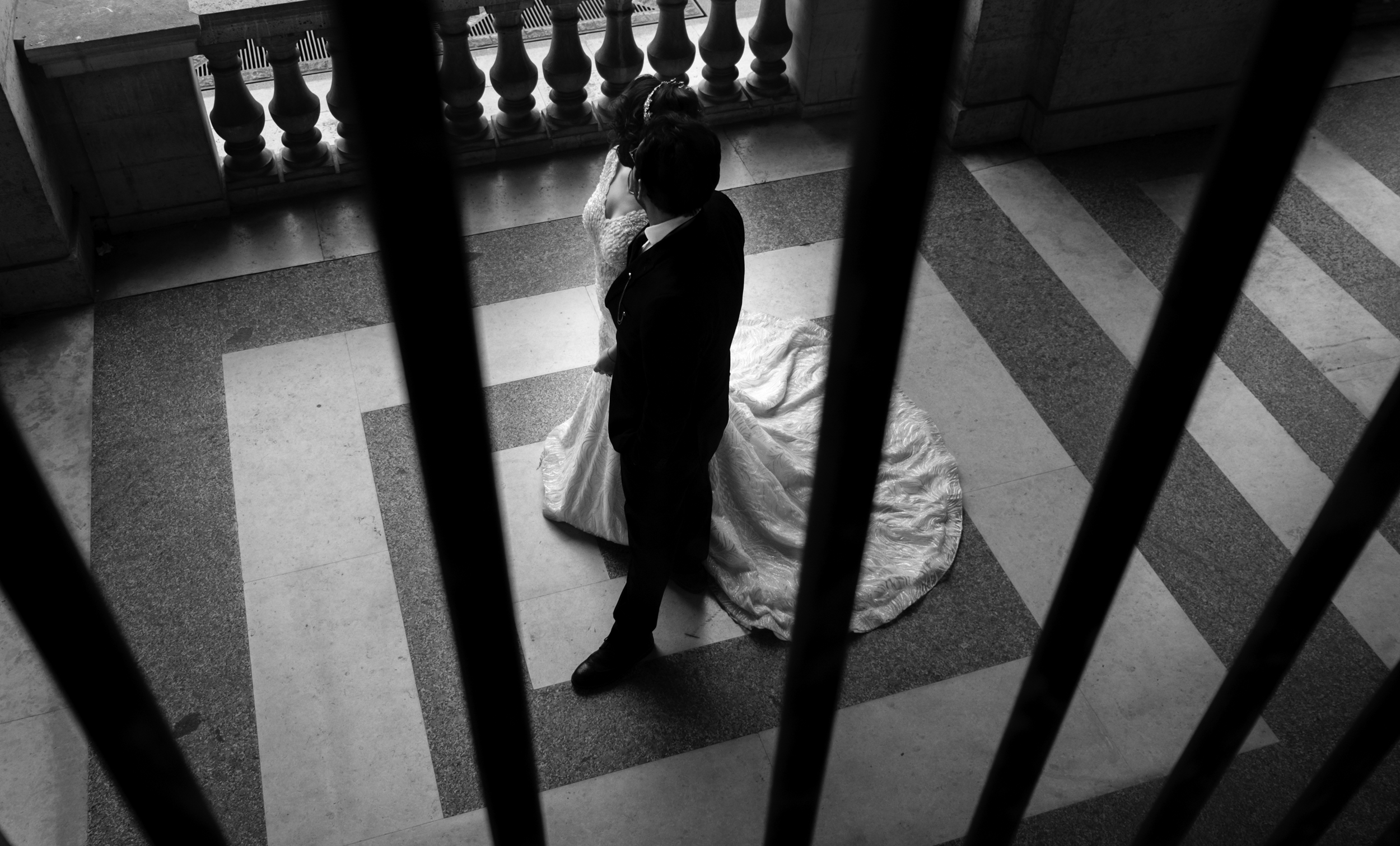 A couple in wedding attire, the woman in a wedding dress and the man in a suit, stand close together on a patterned floor near a balustrade, viewed from above through black bars.