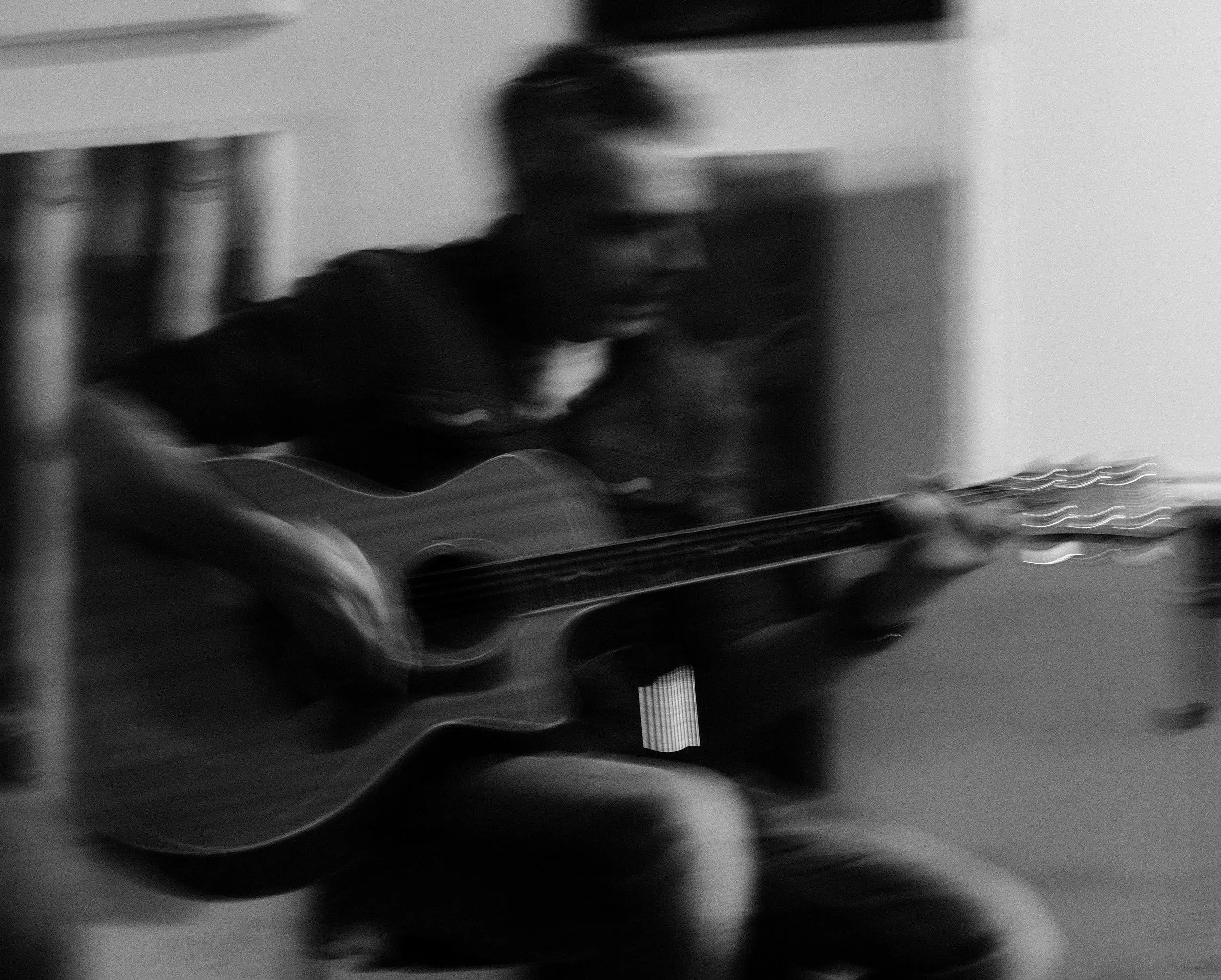 A blurry black and white photo of a person in a dark jacket, possibly wearing glasses, standing near a counter or table with items on it. The background appears to be an indoor space with a wall and some objects.