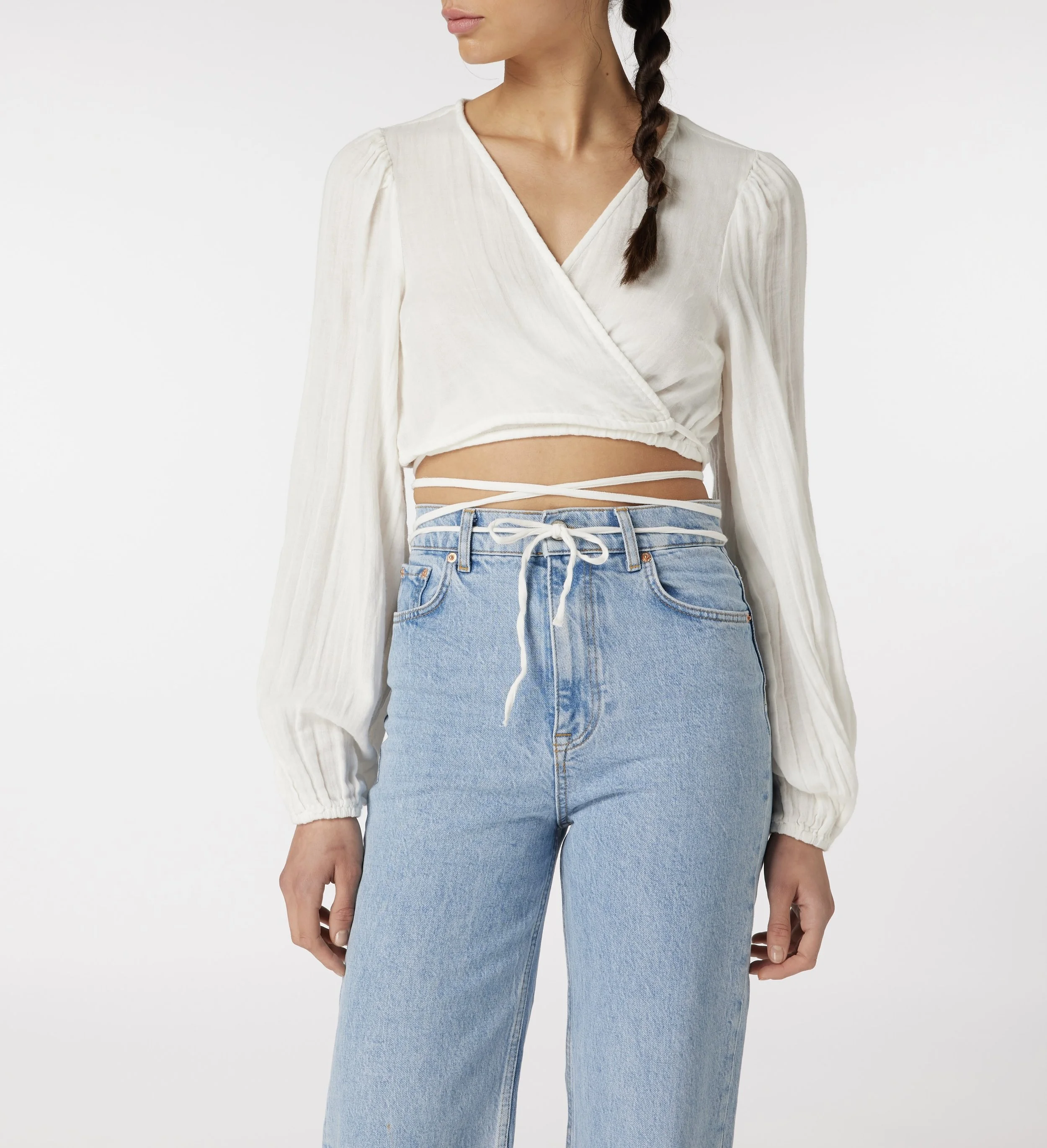 Close-up of a woman wearing a white, long-sleeved, wrap-top with a V-neck and high-waisted light blue jeans, standing against a plain white background.