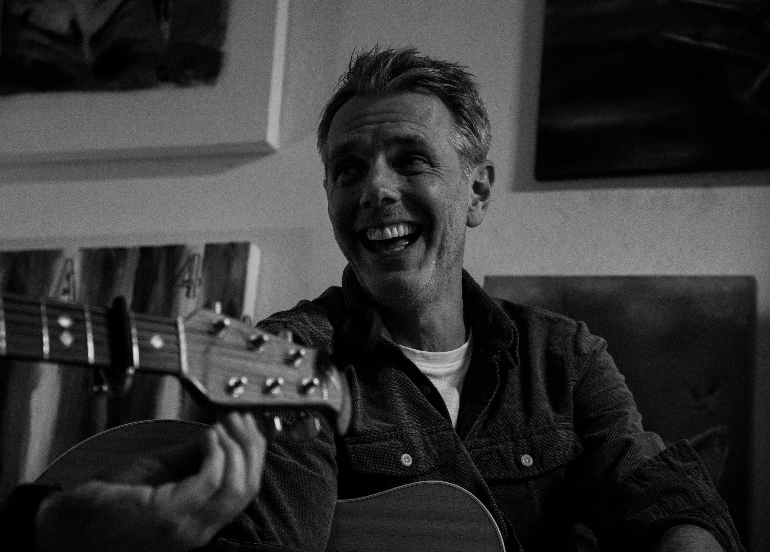 Black and white photo of a man smiling while playing an acoustic guitar indoors. The man has short, styled hair and is wearing a checkered shirt.