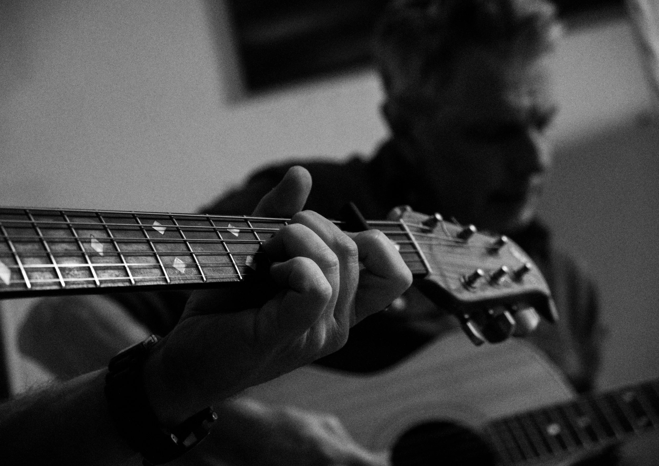 A person playing an acoustic guitar in black and white, with focus on their fingers on the fretboard and their face blurred in the background.