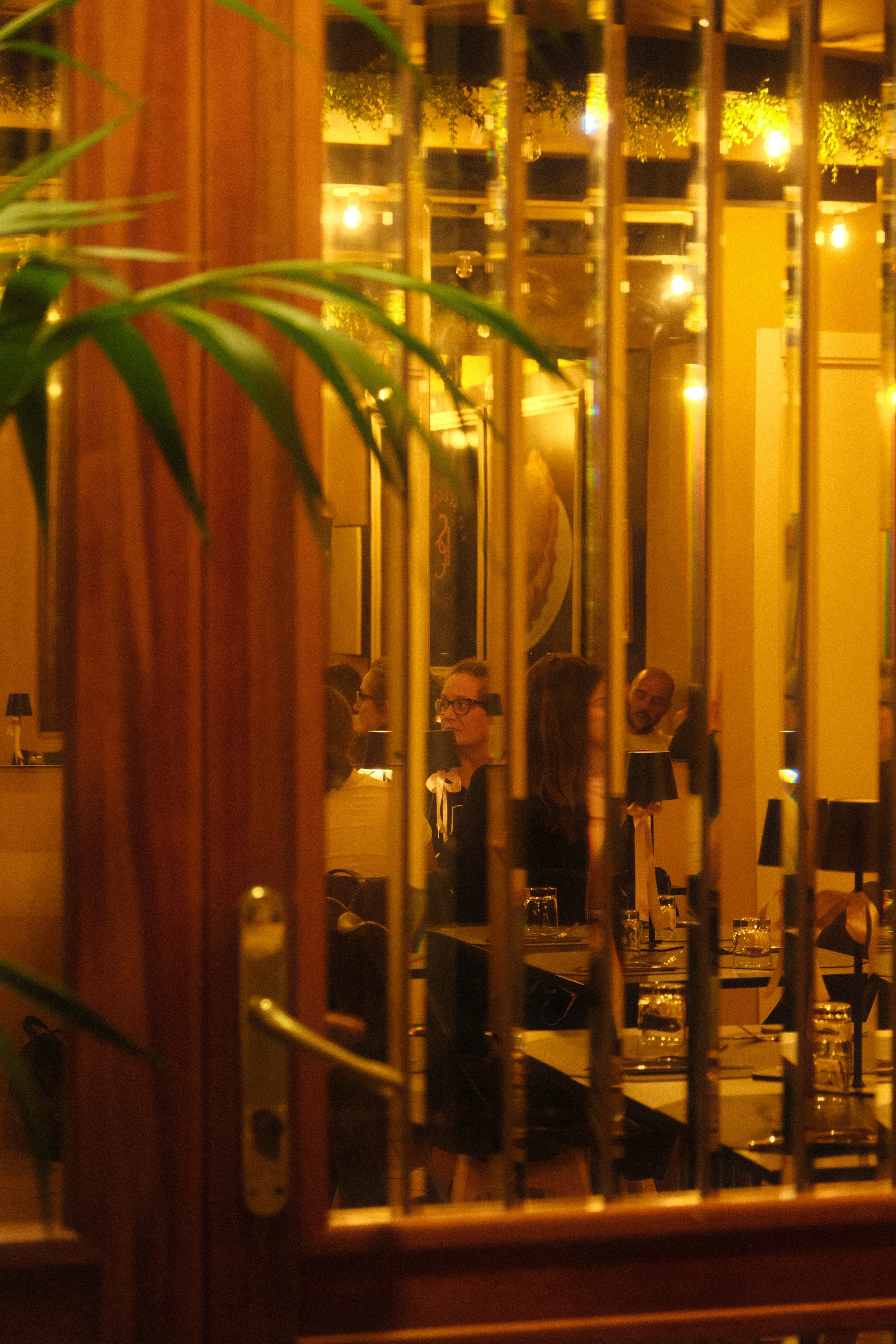 People sitting at tables inside a warmly lit restaurant, viewed through a barred window, with some greenery visible.