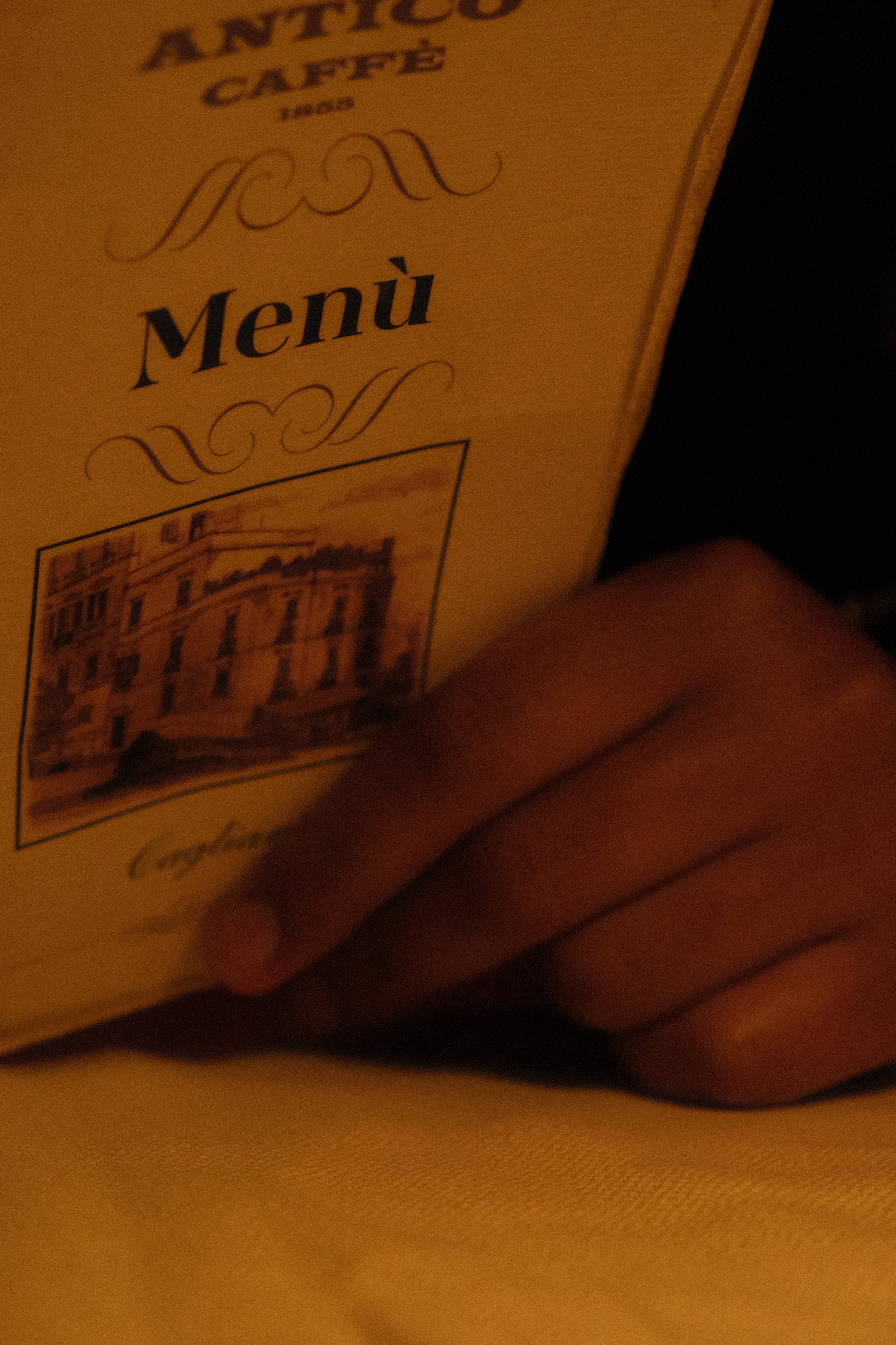 Close-up of a menu cover labeled 'Antico Caffe' with a picture of a building underneath. A person's hand is holding the menu, and the background is dark.