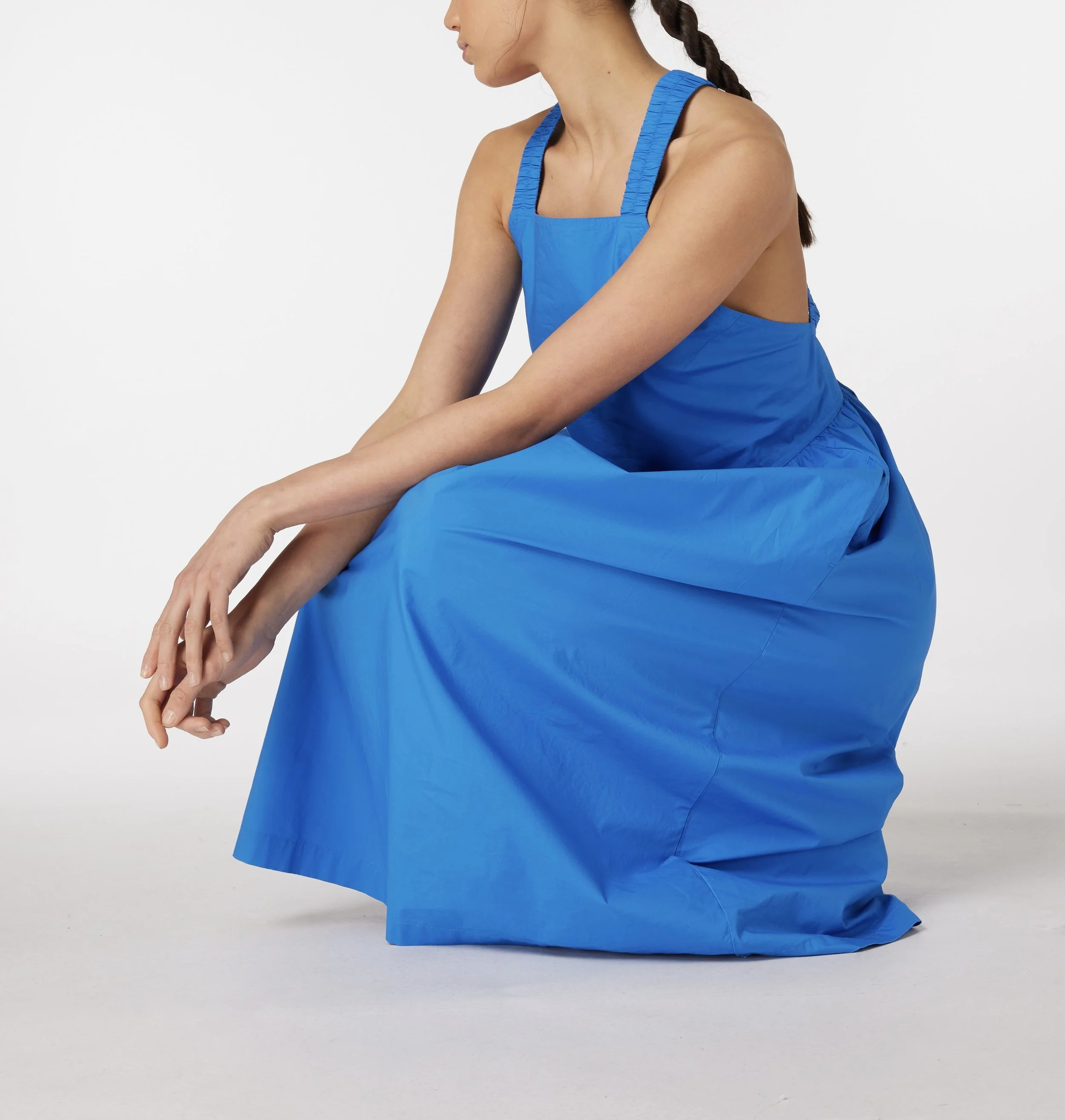 A woman in a blue dress is kneeling on a white surface against a plain white background, with her arms resting on her lap and her head turned to the side.