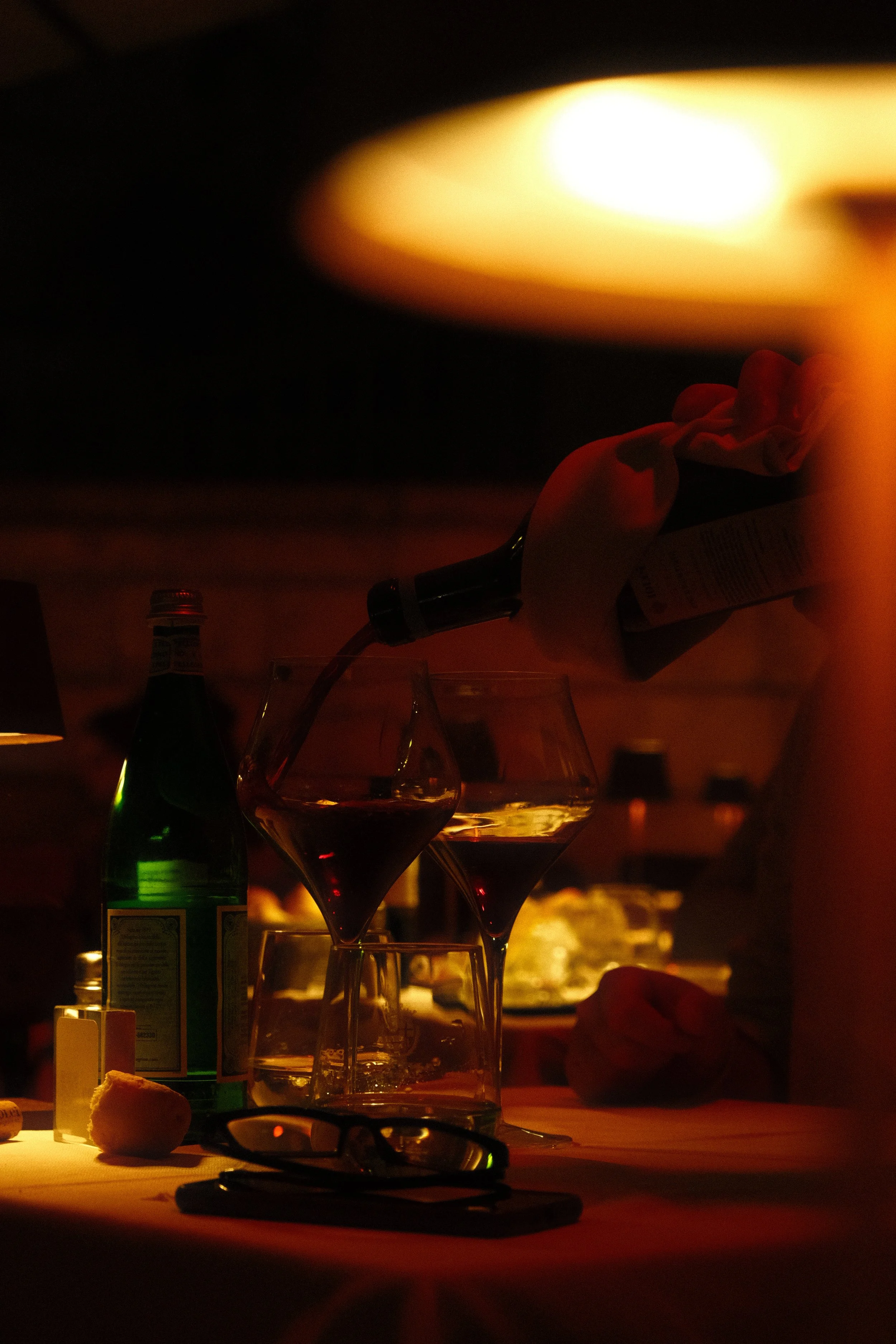 A dimly lit scene with a hand holding a newspaper, liquor bottles, wine glasses, eyeglasses, and a small loaf of bread on a table. There is warm lighting and a blurred background.