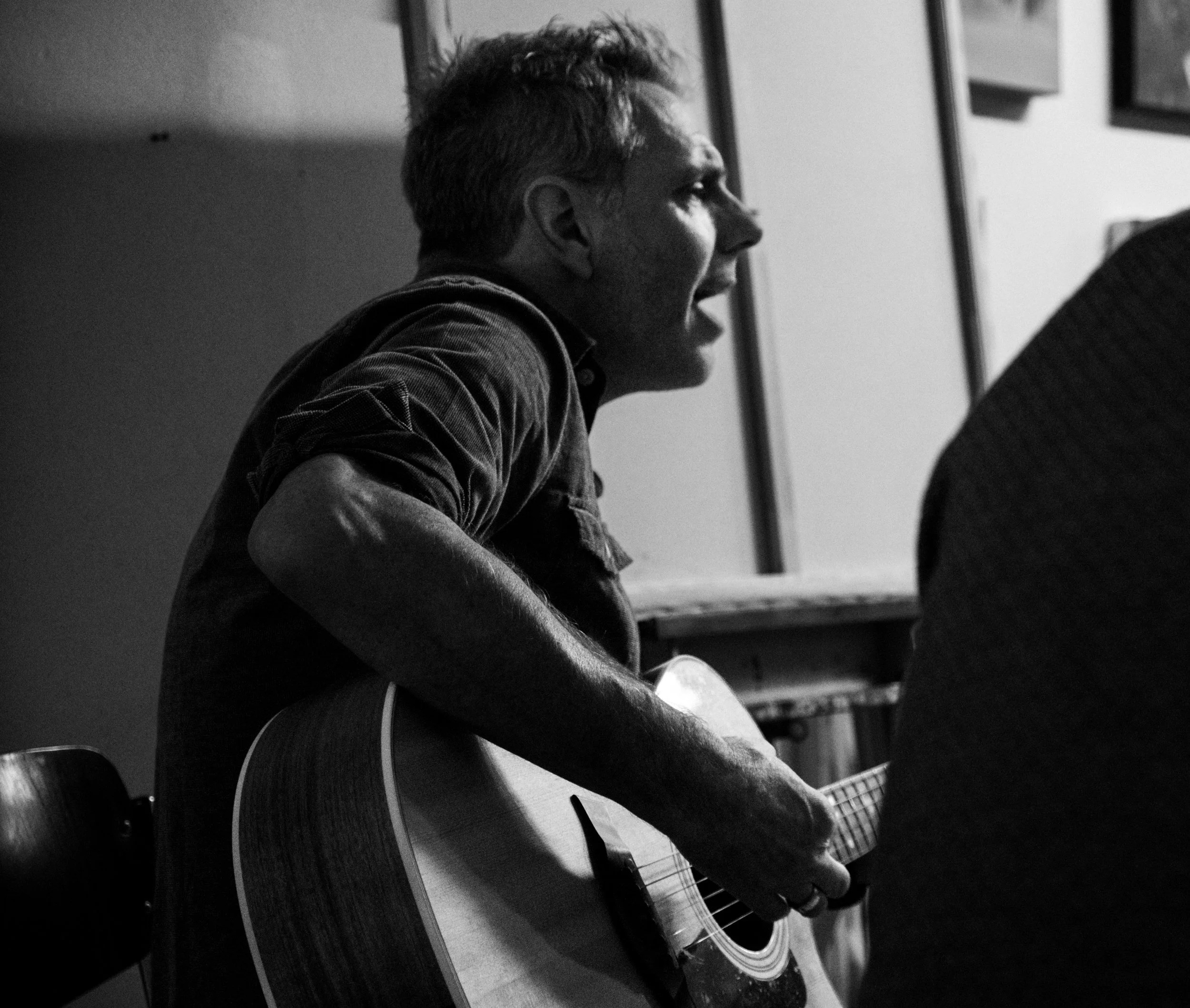 Black and white photo of a man singing and playing an acoustic guitar, sitting on a chair inside a room with framed pictures on the wall.