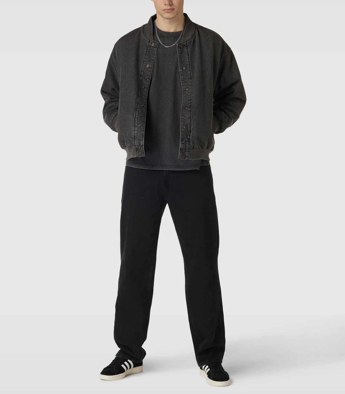A person wearing a black denim jacket, black t-shirt, black pants, and black sneakers with white stripes stands against a plain gray background.