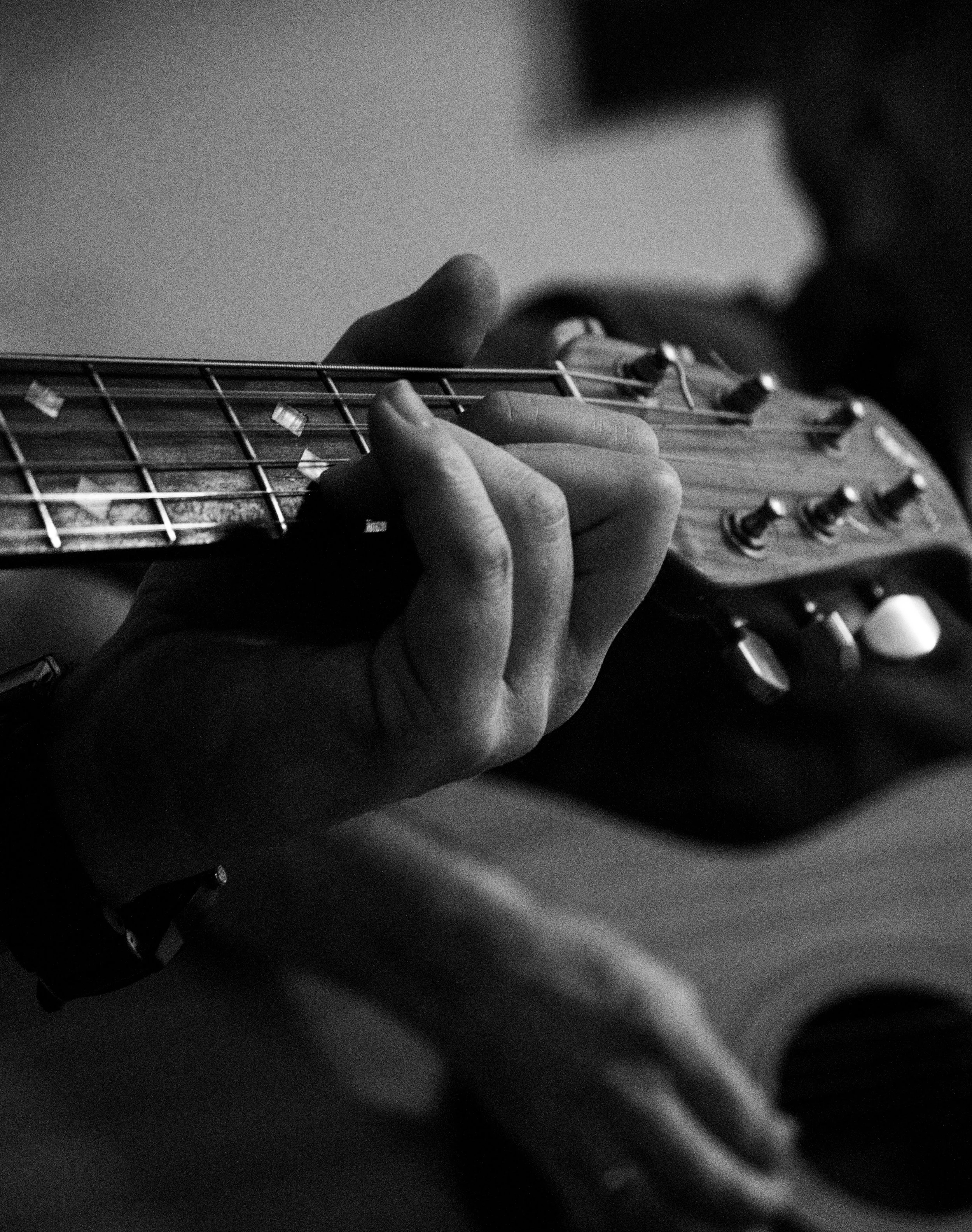 Close-up of a person playing an electric guitar in black and white, focusing on the hand pressing on the fretboard and the tuning pegs.