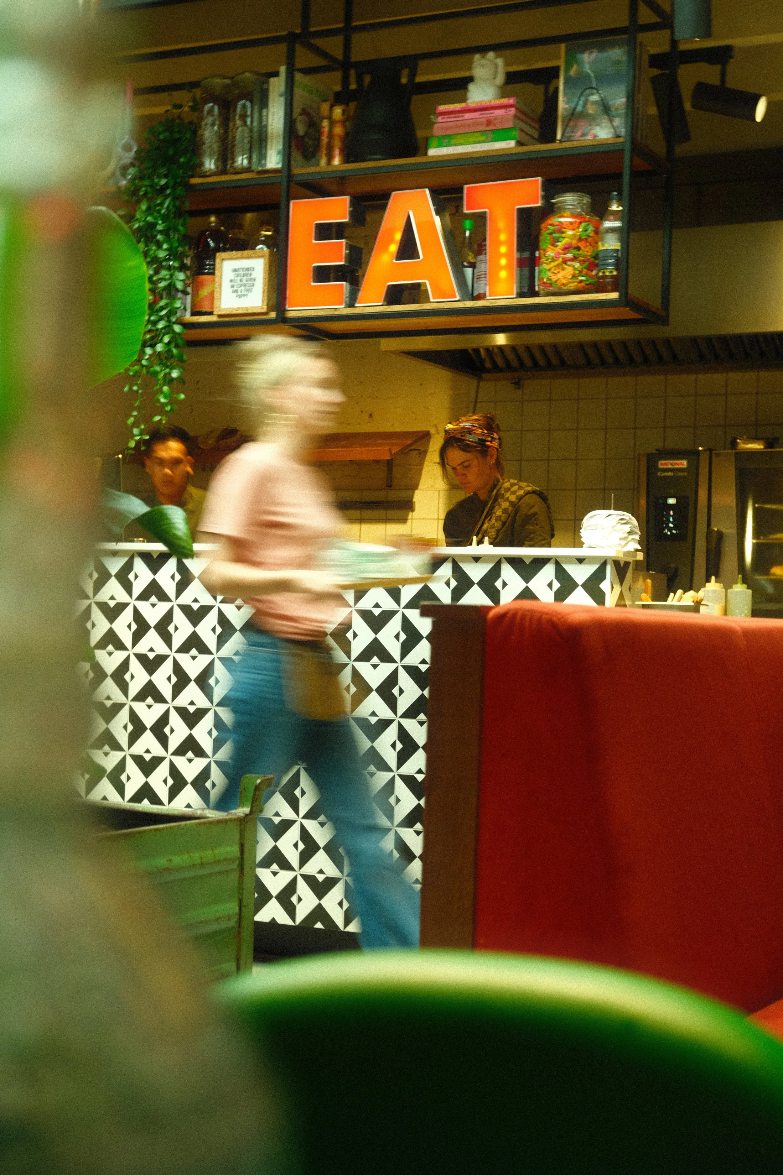 Inside a restaurant with a kitchen and a countertop. There are decorative shelves with books, jars, and ornaments. A large illuminated sign spelling 'EAT' is on the shelf. Several people are in the space, some blurred in motion, one woman is working 