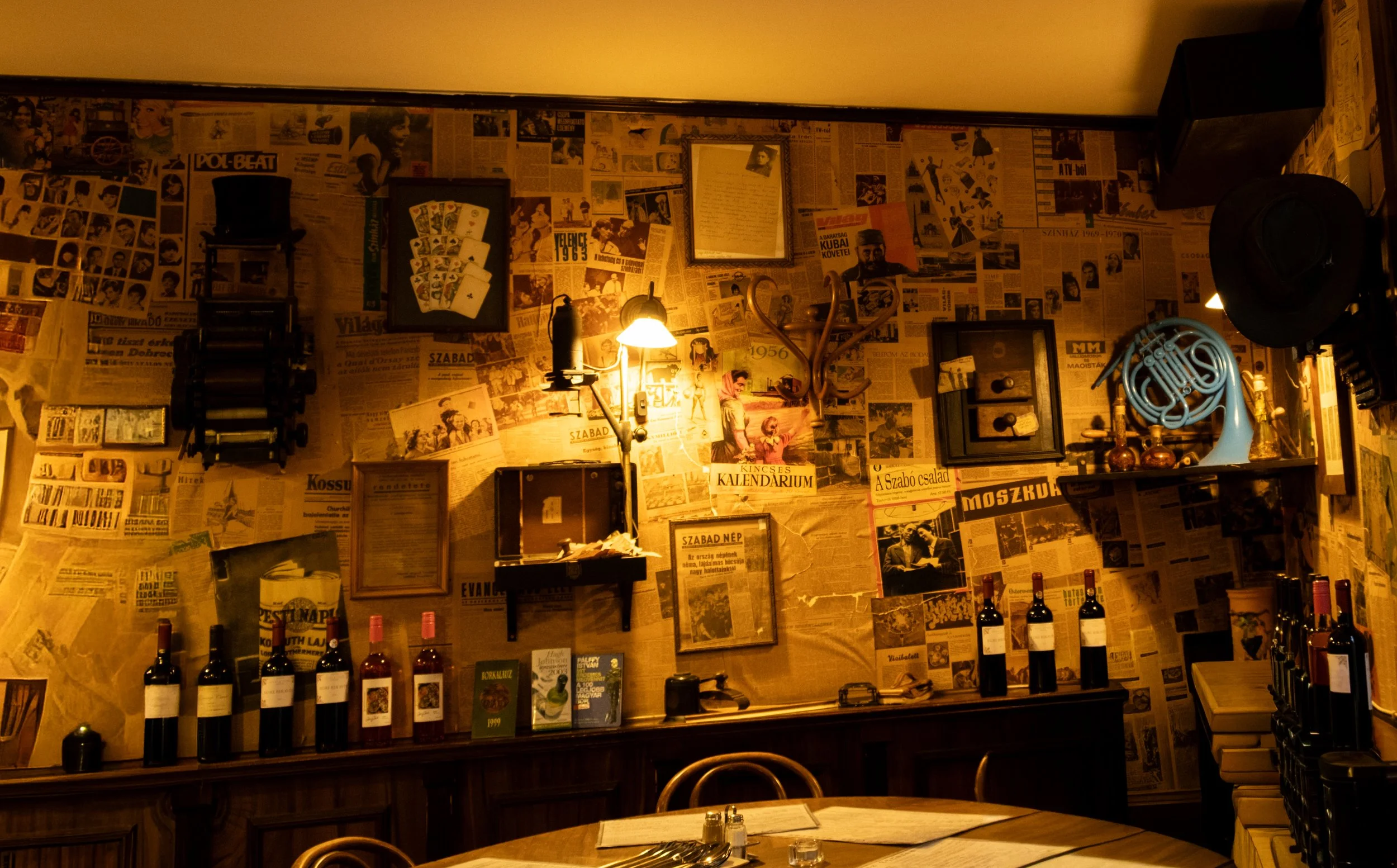 Interior of a restaurant or bar with a wall covered in newspaper clippings, framed photos, a lamp, and various decorative objects, including wine bottles on a shelf.