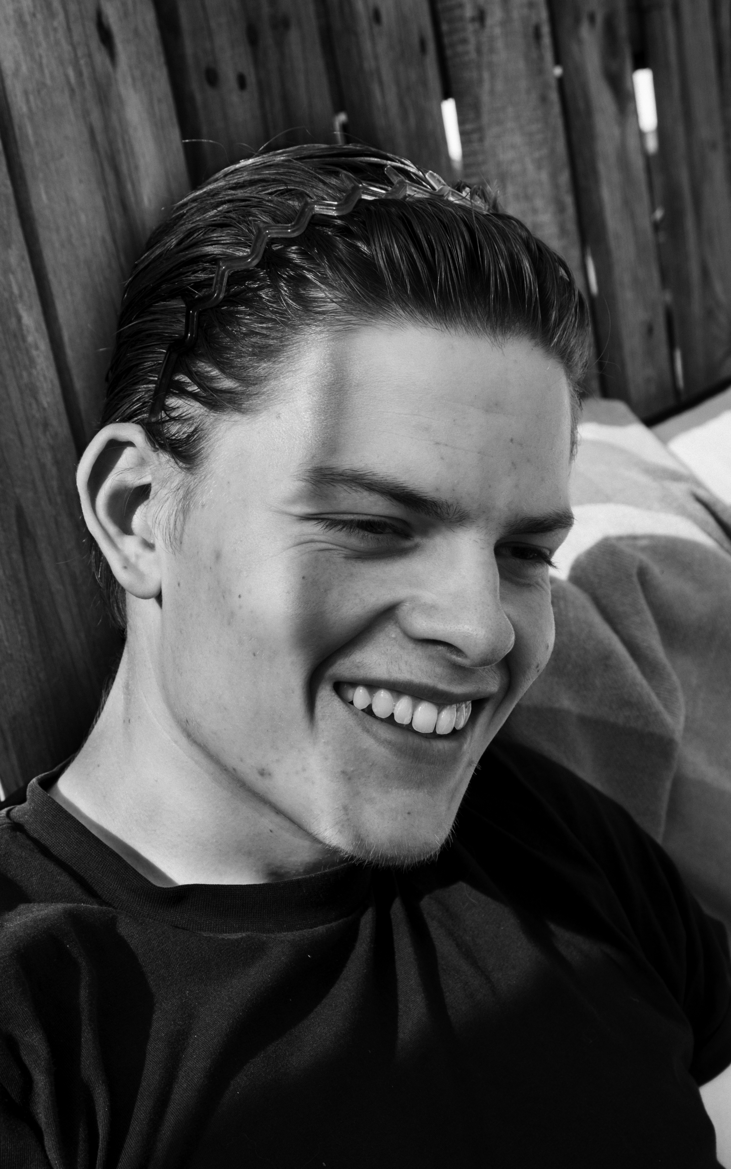 Close-up of a smiling young man with slicked-back hair and a wavy hair clip, sitting against a wooden wall.