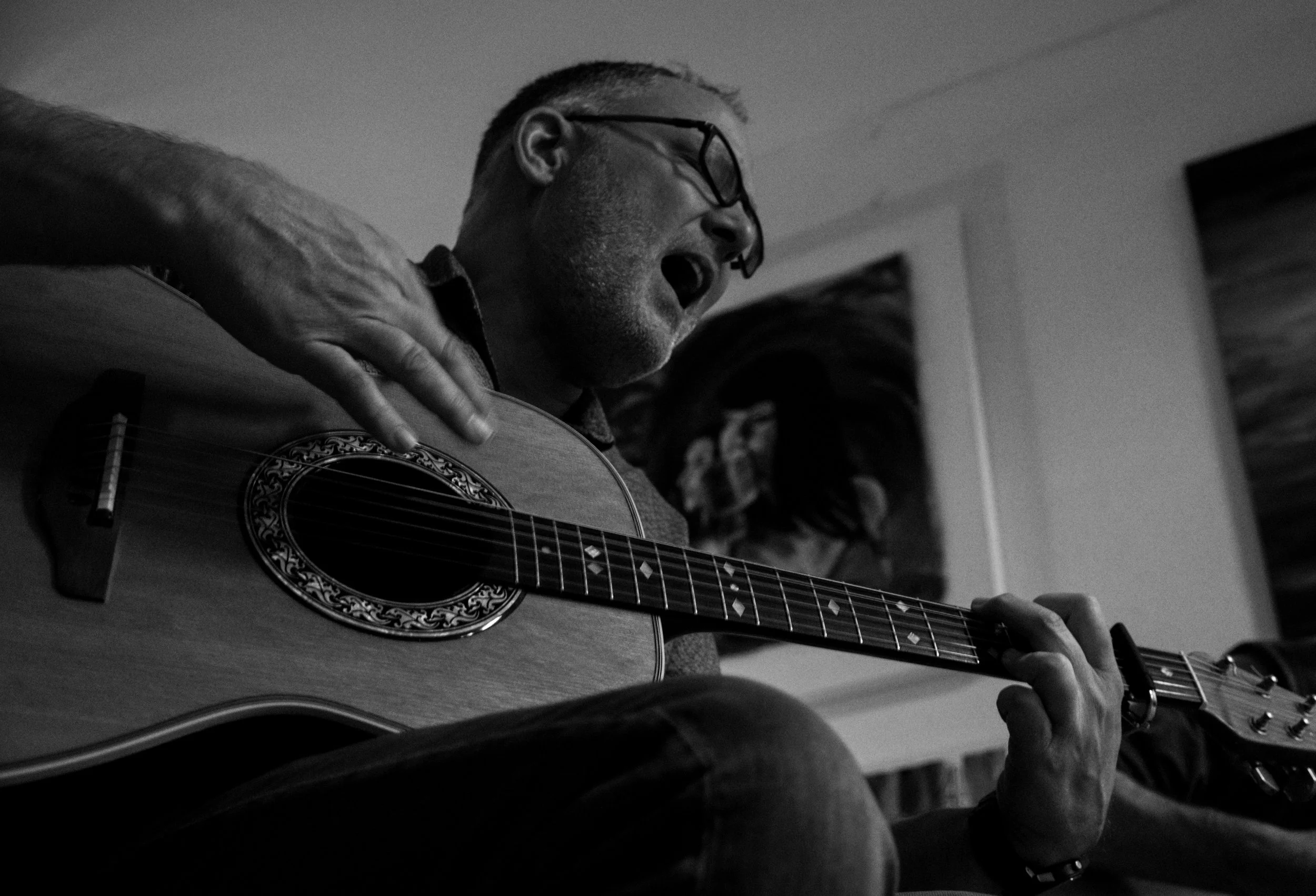 A man wearing glasses playing an acoustic guitar in a room, captured in black and white.