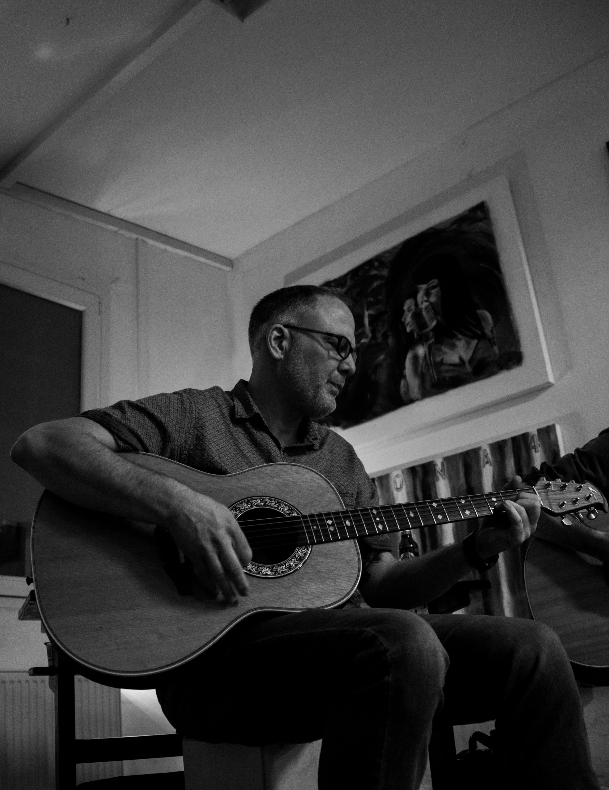 A man with glasses playing an acoustic guitar indoors, sitting in front of a wall with a painting of people.
