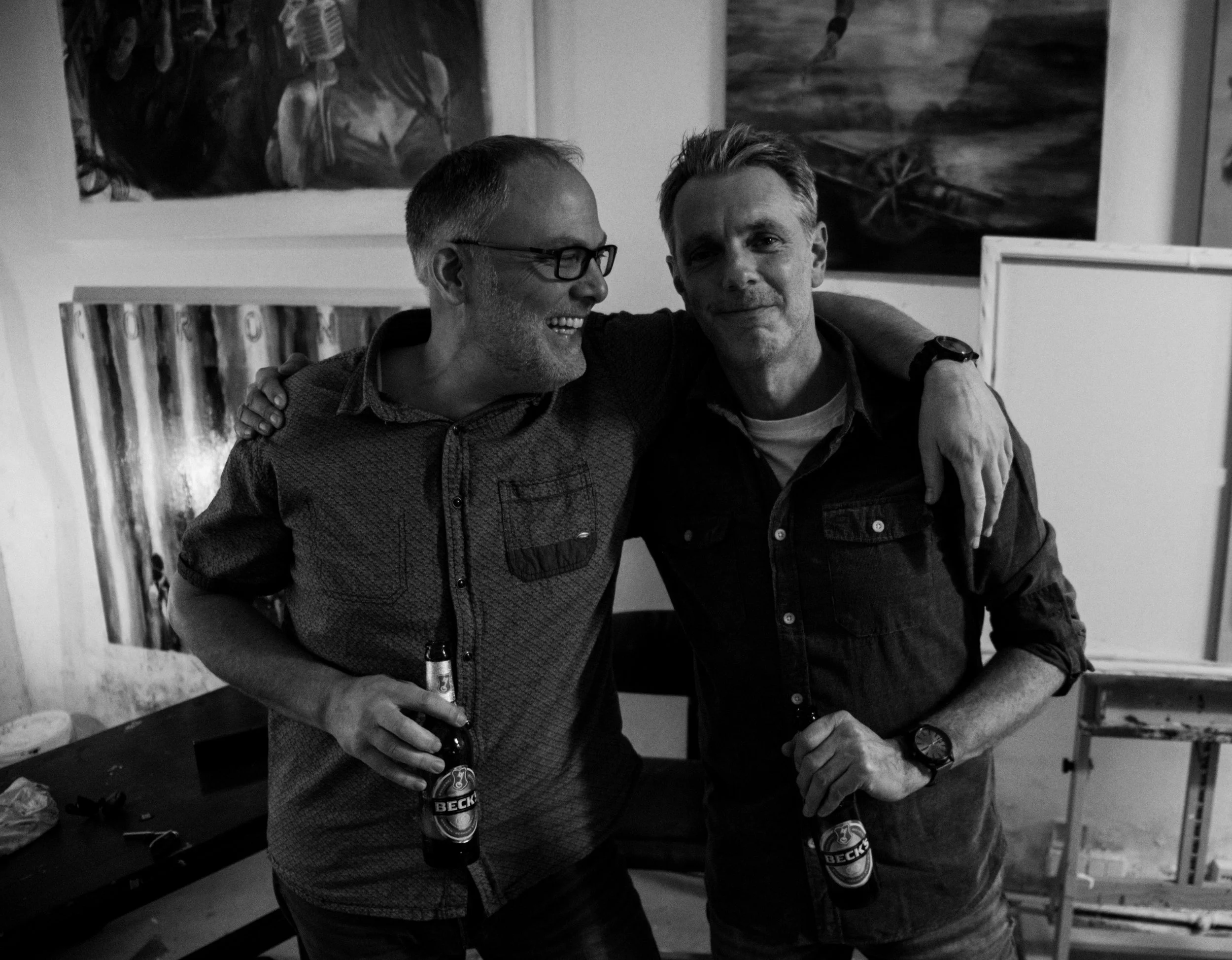 Two men sharing a moment at a social gathering, smiling and holding bottled beverages, one with glasses, the other without, standing in a room with artwork on the wall.