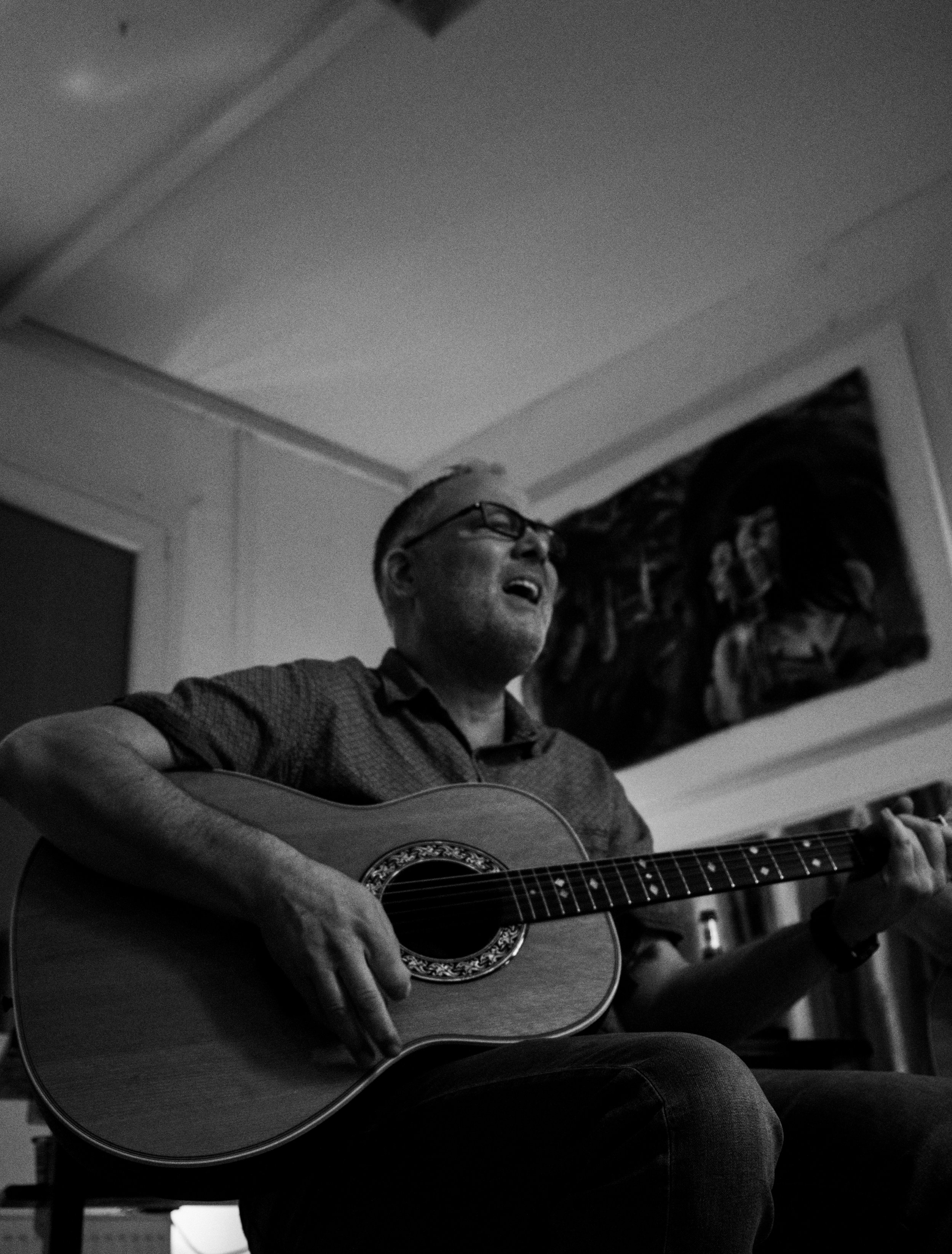 A man wearing glasses and a short-sleeved striped shirt is playing an acoustic guitar in a room with decorated walls. He appears to be singing or enjoying the music.
