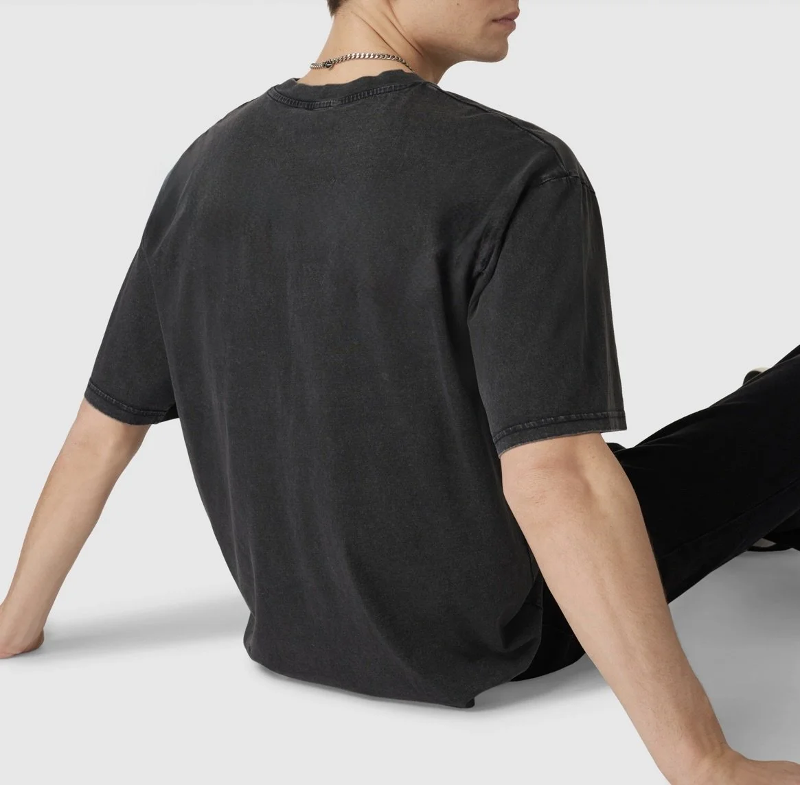 A person sitting on the floor wearing a black T-shirt and black pants, with a silver chain necklace, against a plain gray background.