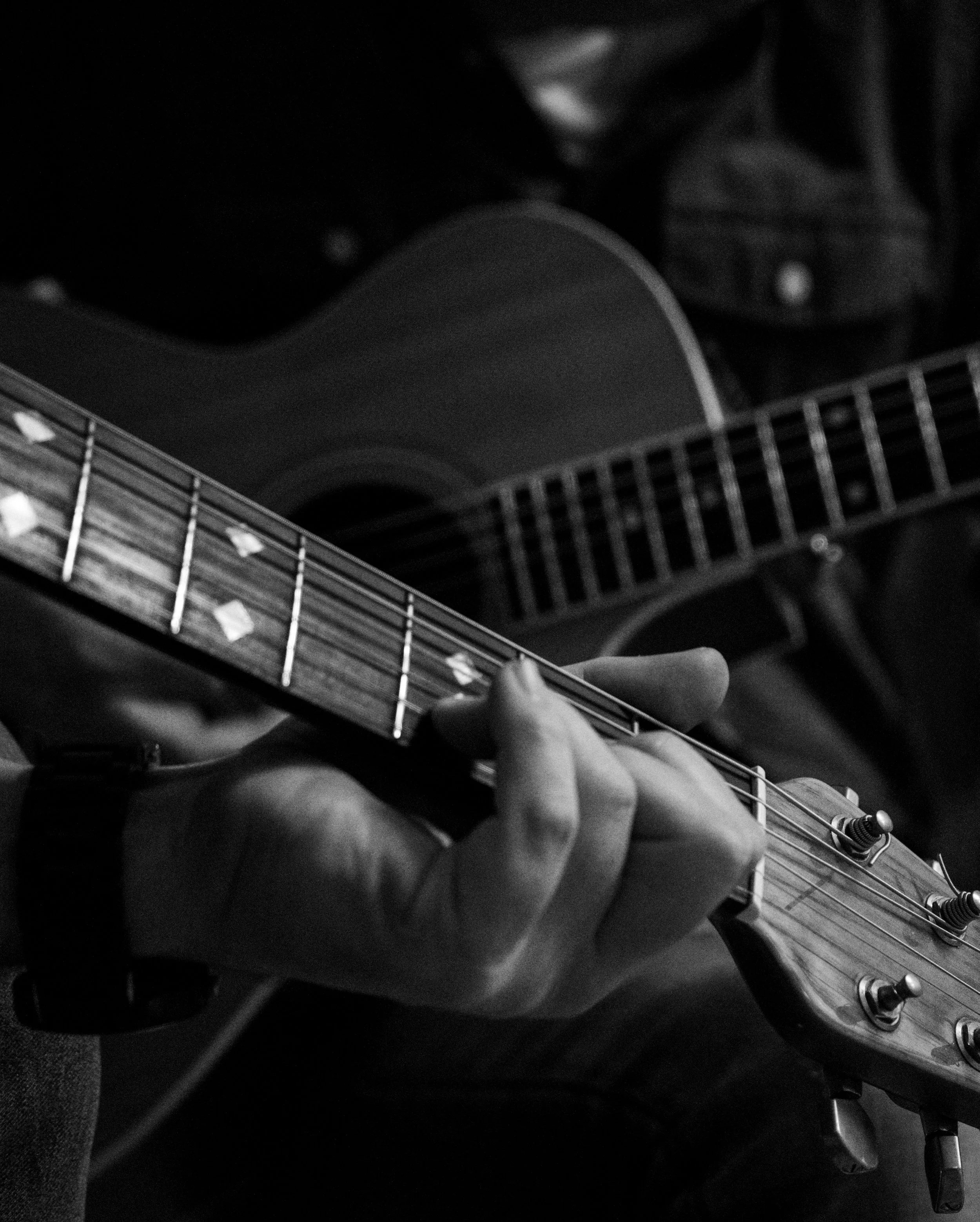 Close-up black and white photo of a person playing an acoustic guitar, focusing on the guitar neck and the person's hand pressing down on the strings.
