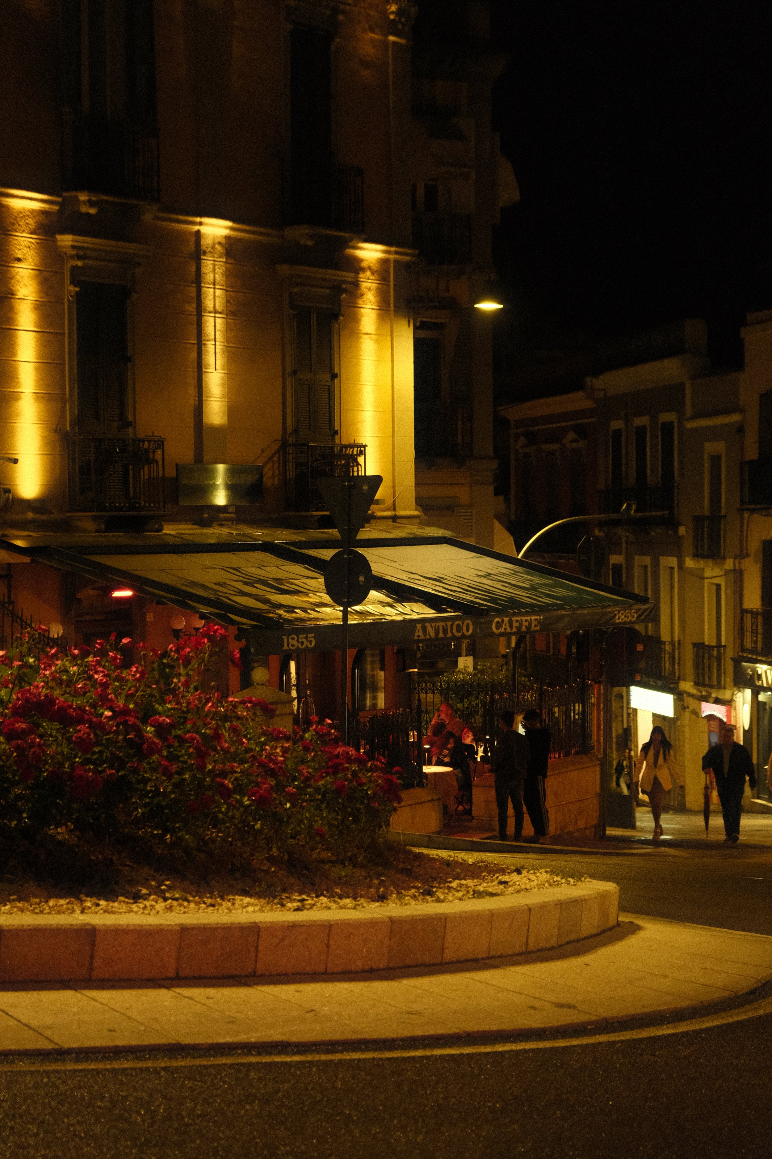 Night scene of a city street with a building lit by yellow lights, people walking on the sidewalk, and a storefront with a sign that reads 'ANTICO CAFE'. There are also some flowers in the foreground and a streetlamp hanging overhead.