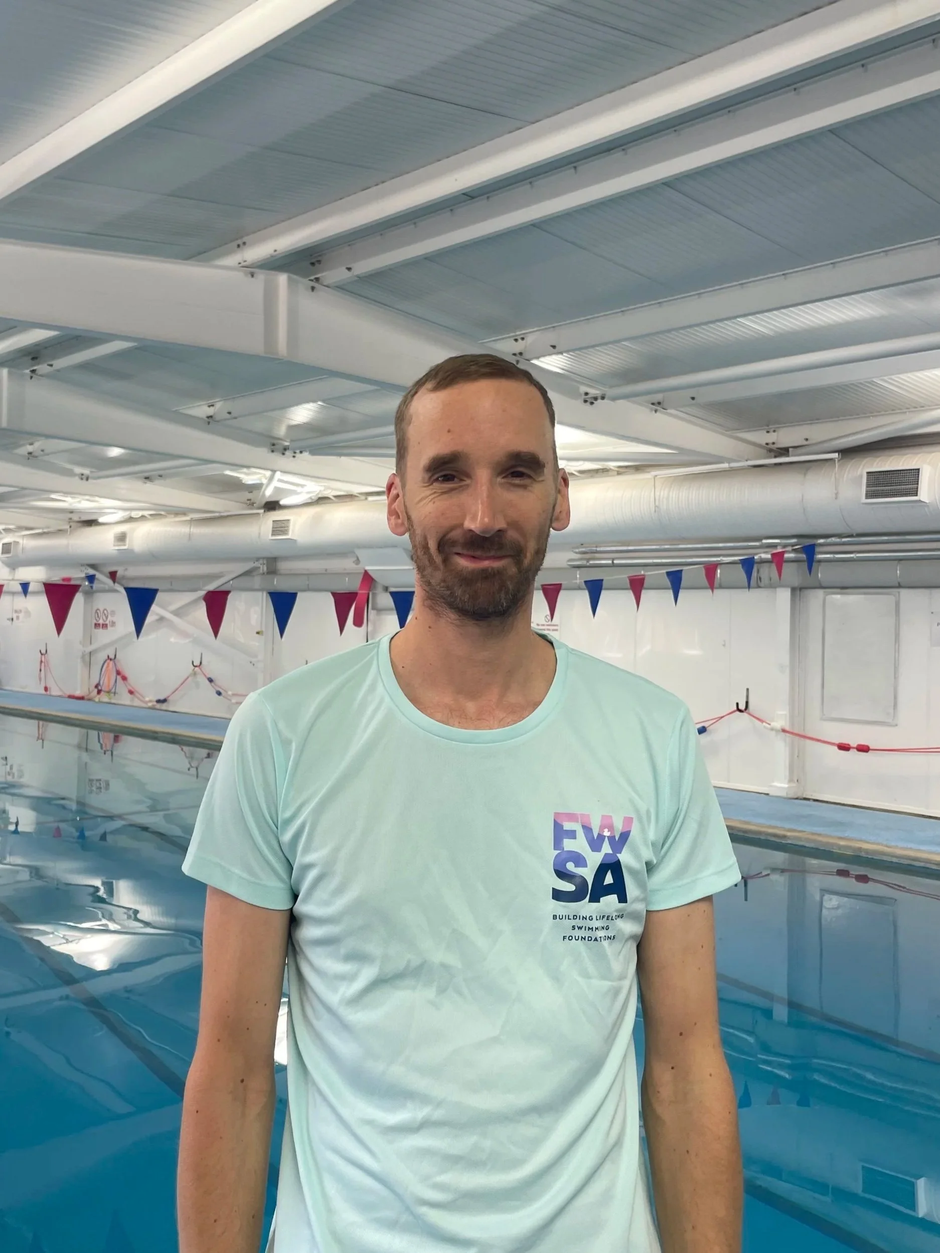 A man standing next to a swimming pool inside an indoor swimming facility, wearing a light-colored T-shirt with 'FVSA' logo.
