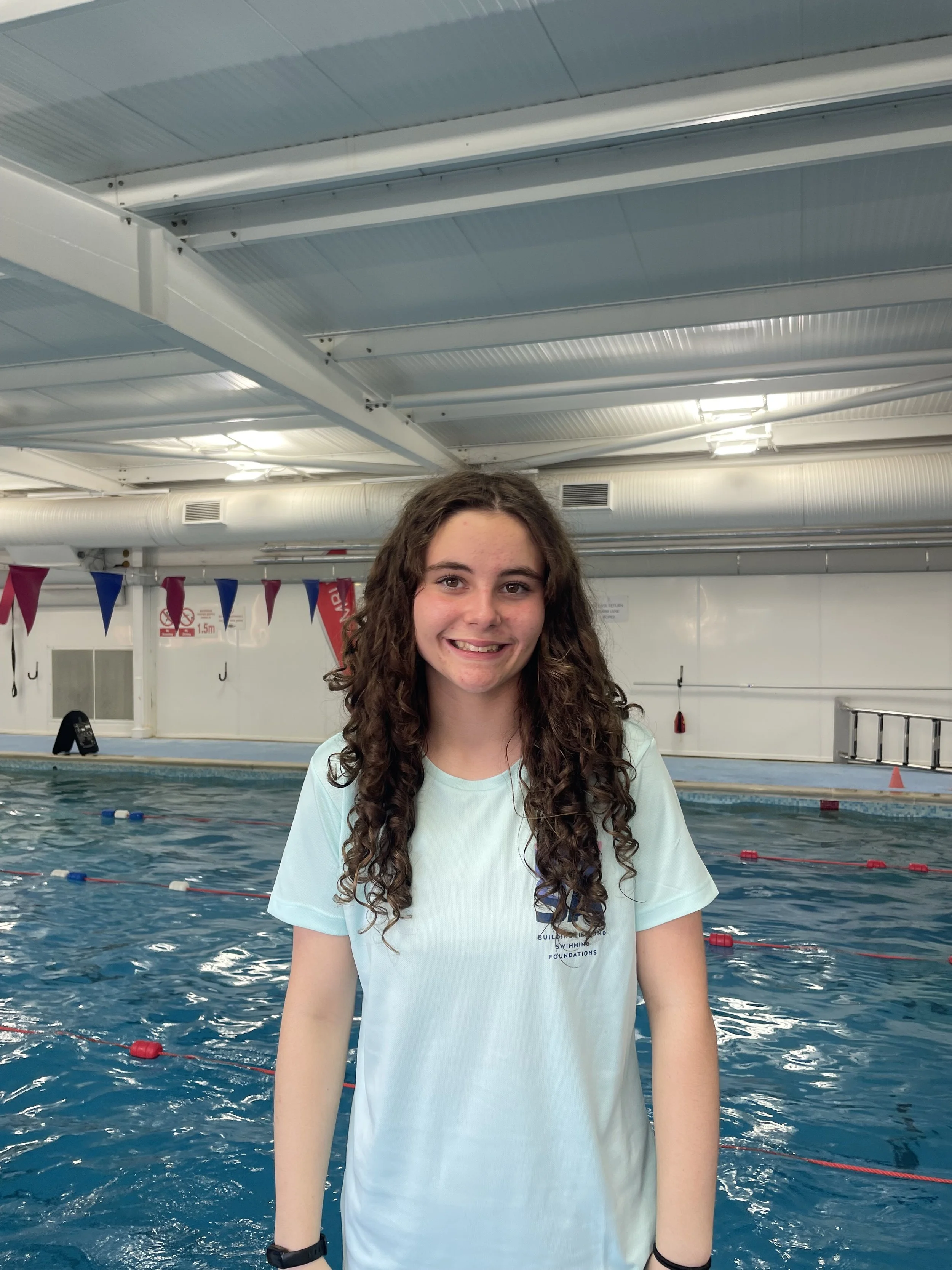 A young woman with long, curly brown hair smiling at the camera, standing by an indoor swimming pool with lanes marked by red and blue lane dividers, and a white wall with equipment and flags in the background.