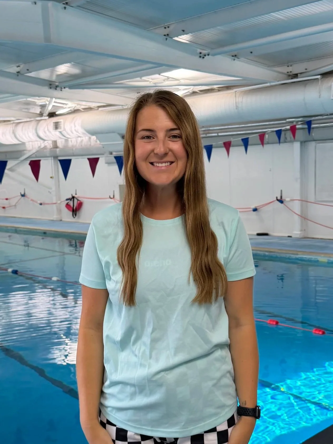 A smiling woman with long brown hair standing at the edge of an indoor swimming pool, with blue and pink flags hanging above and pool lanes in the background.