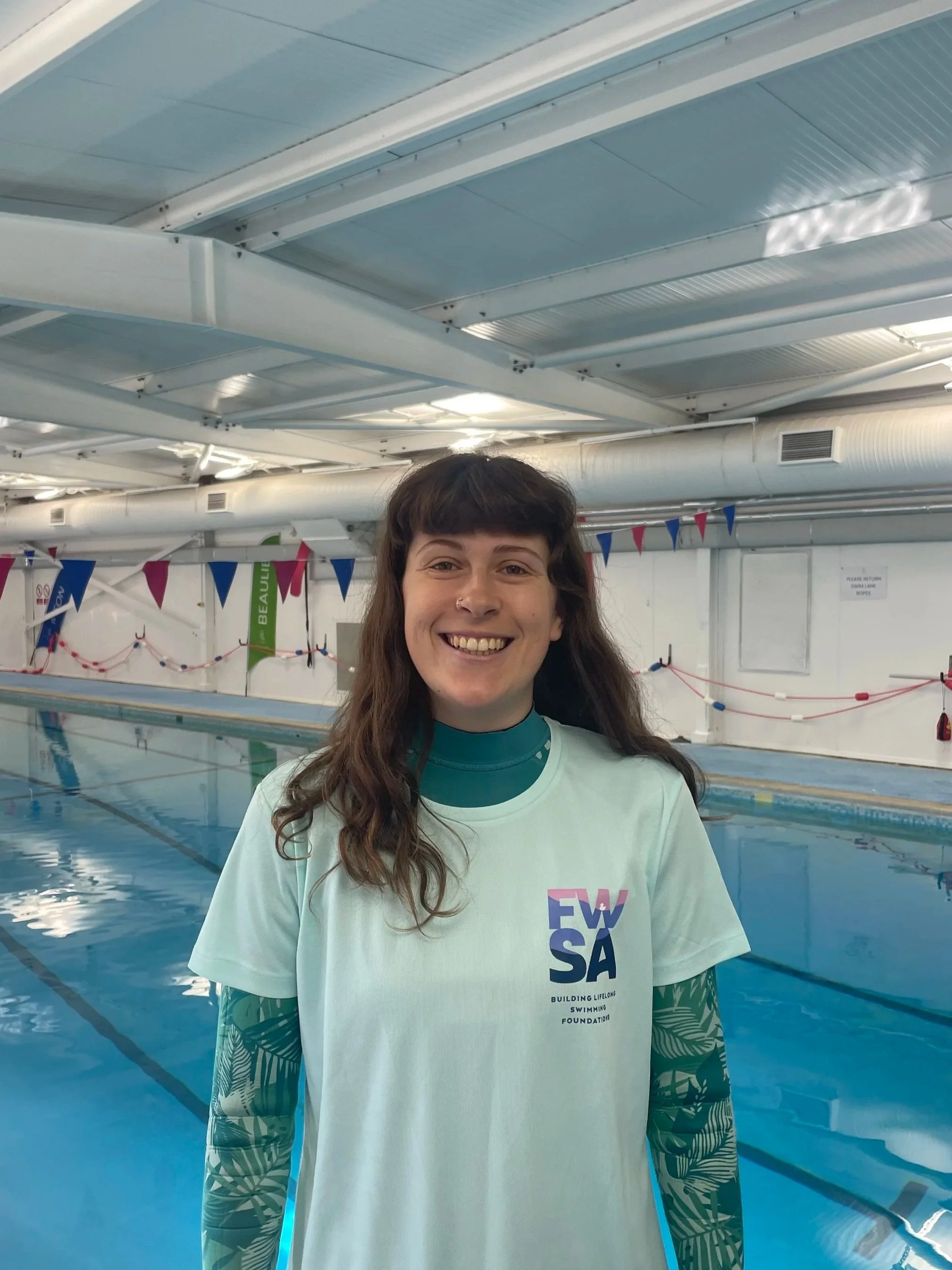 Young woman with long wavy brown hair smiling at the camera, wearing a white t-shirt with text, at an indoor swimming pool.