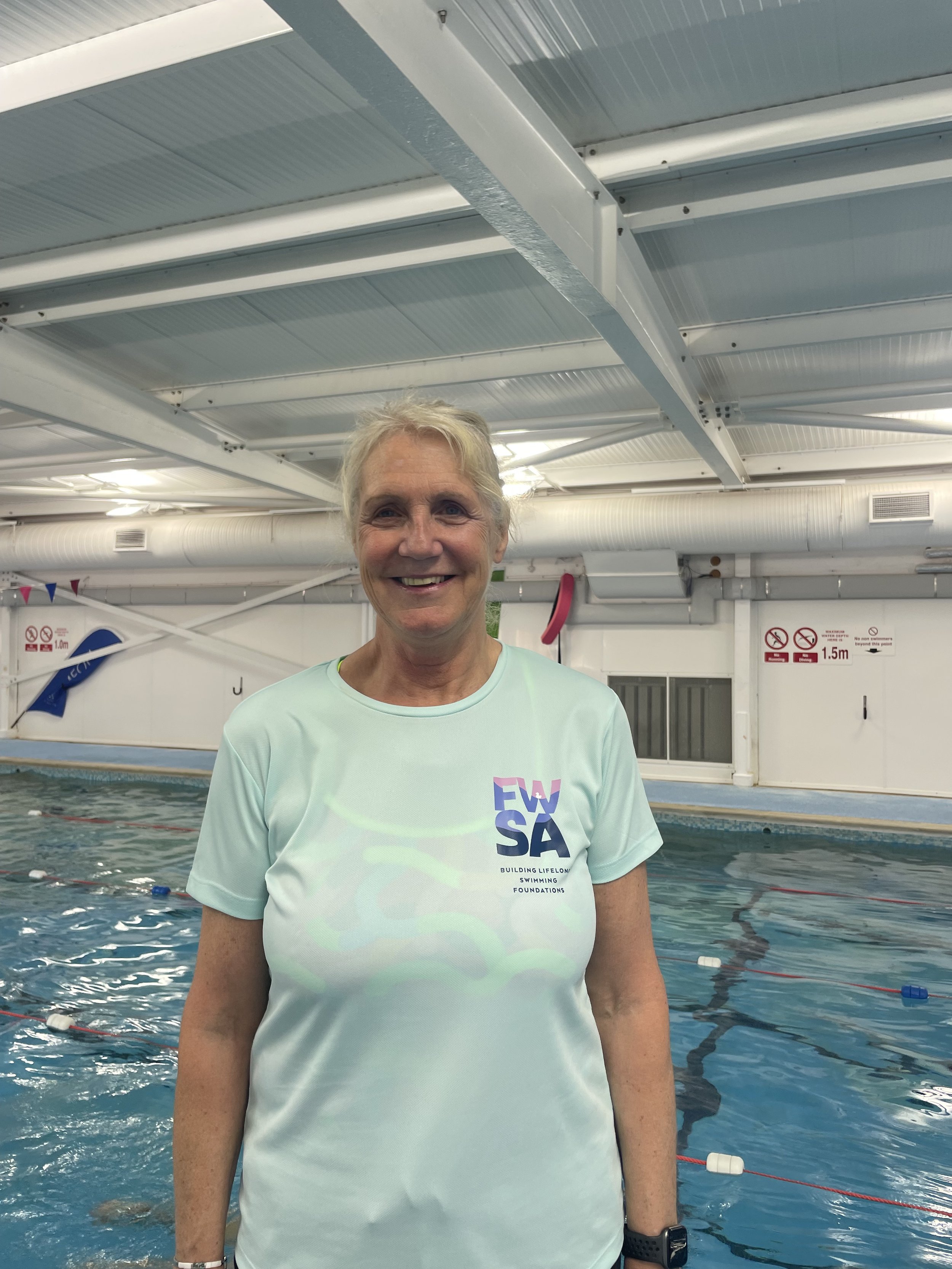 A smiling older woman with short blonde hair standing in a swimming pool, wearing a light green T-shirt with 'ESWSA' and 'Building Lifelong Swimming Foundations' printed on it. The indoor swimming pool has a white metal ceiling, ventilation ducts, and safety signs on the wall.