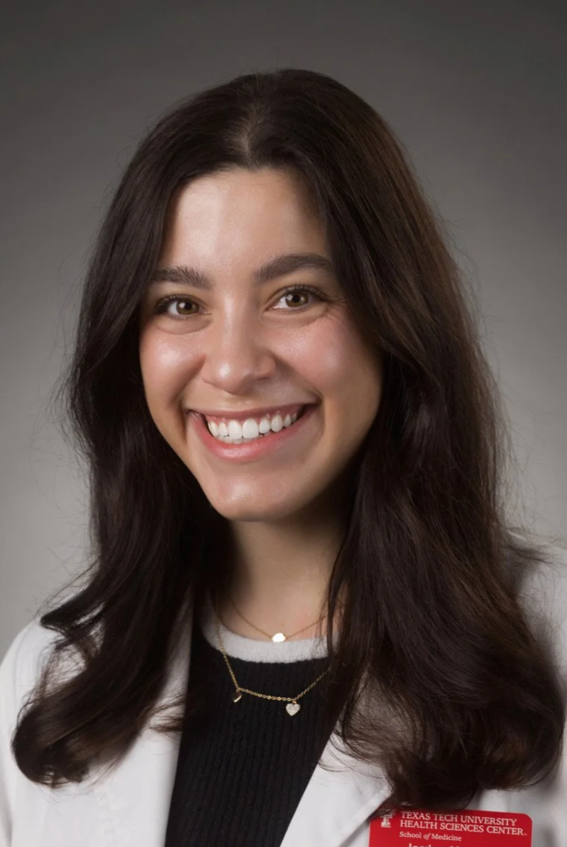 Portrait of a young woman with long dark hair smiling, wearing a white lab coat with a medical school badge, black top, and gold jewelry, against a gray background.
