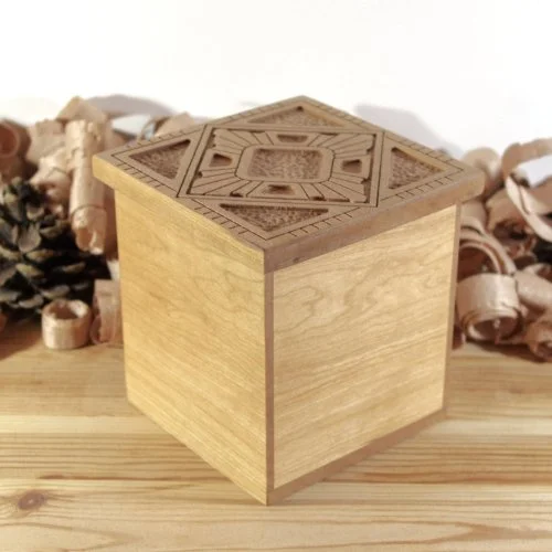 A wooden cube with carved geometric patterns on its top surface, placed on a wooden table with wood shavings and wood chips in the background.