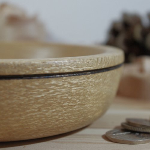 Close-up of a light-colored wooden bowl with a dark rim, placed on a wooden surface with some coins nearby.