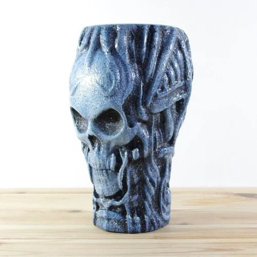 A wooden skull-shaped vase with a textured surface and dark blue and black colouring, sitting on a wooden surface.