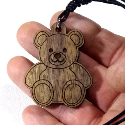 Wooden teddy bear pendant with black cord, held between fingers.