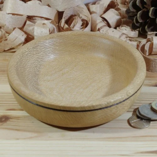 A light-colored wooden bowl on a wooden surface, with white wood shavings and coins nearby, and a pinecone in the background.
