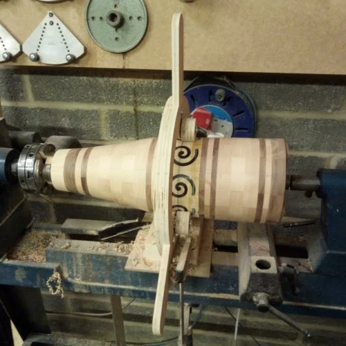 A woodworking lathe with a partially turned wooden piece mounted on it, holding a small wooden disc with spiral patterns, in a workshop setting.