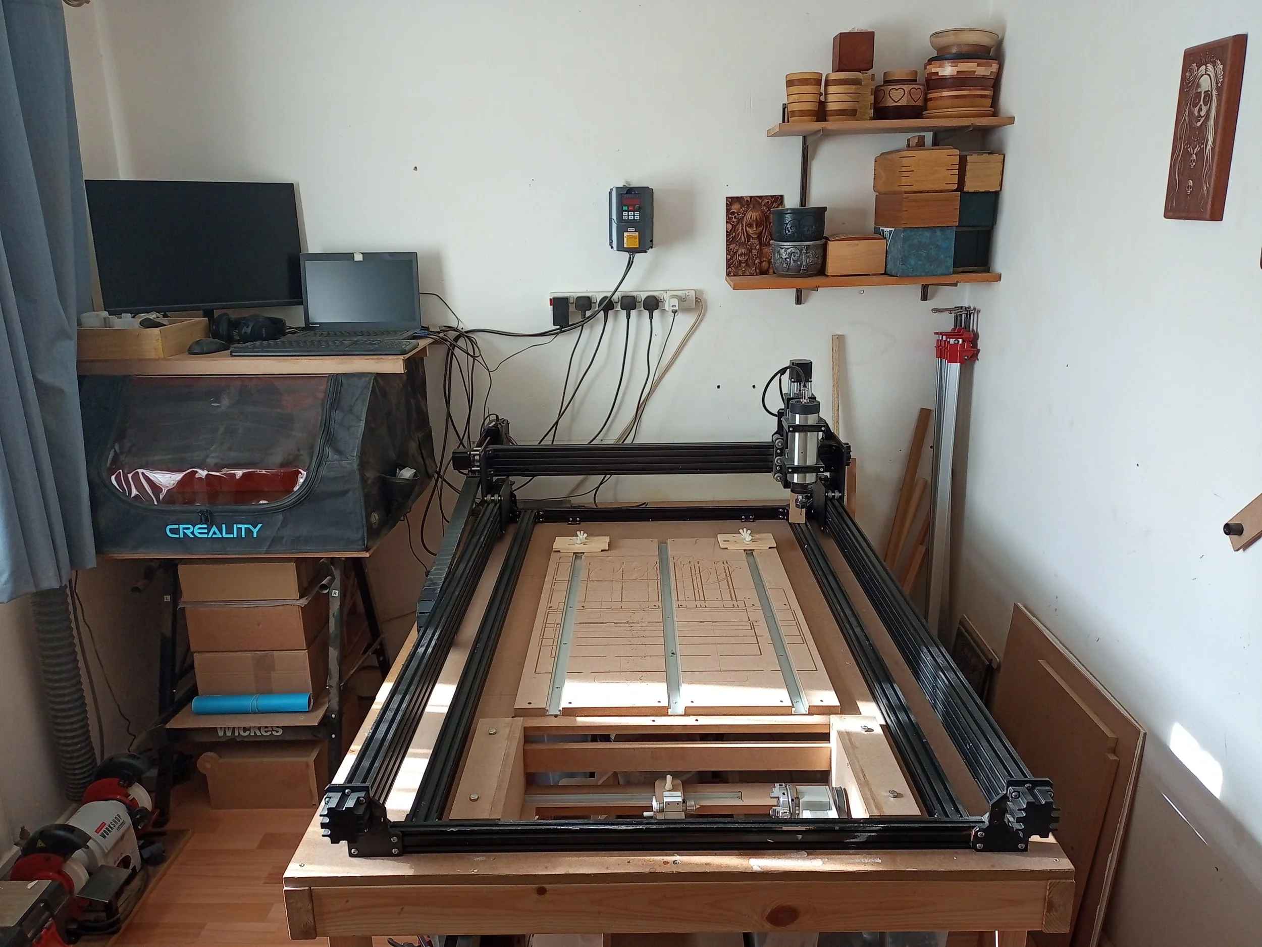 A woodworking CNC machine in a workshop, with wooden panels and tools nearby, and a computer setup on a desk to the left.