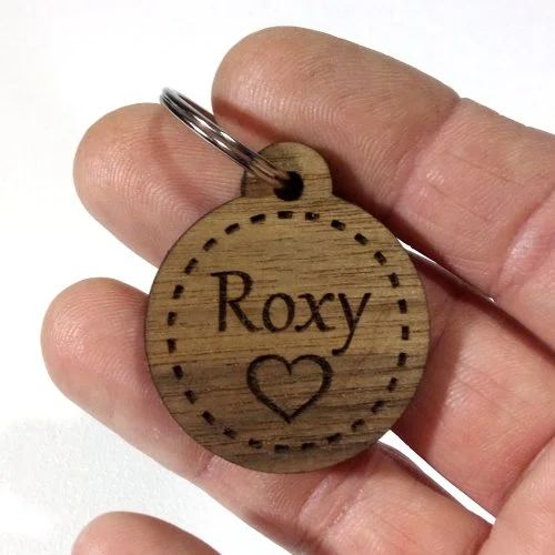 A small round wooden pet tag with the name "Roxy" and a heart symbol, held between fingers.