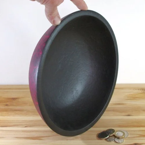 A hand holding a black and red bowl with coins on a wooden surface.