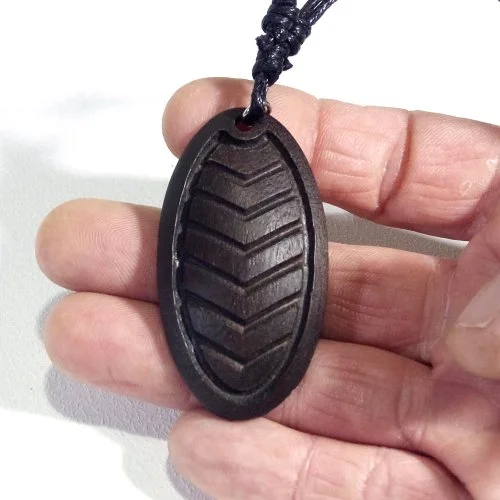 Black oval-shaped pendant with a raised chevron pattern, hanging from a black cord, held by a person's fingers.