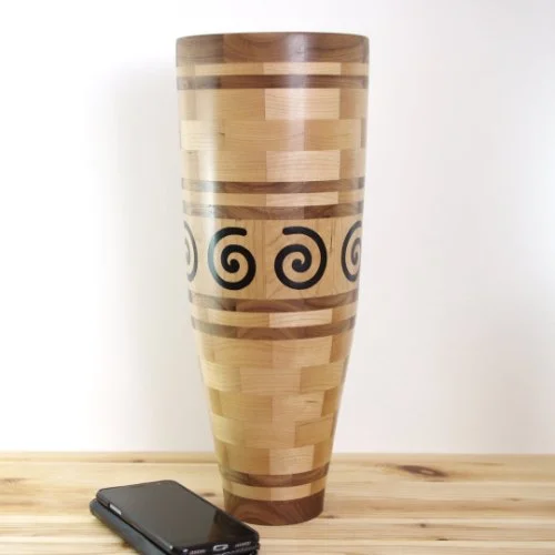 Tall wooden segmented vase with decorative black epoxy spirals on it, resting on a wooden surface with a smartphone nearby.