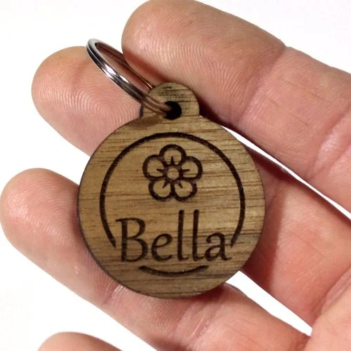 A hand holding a wooden pet tag with a flower engraving and the name Bella.