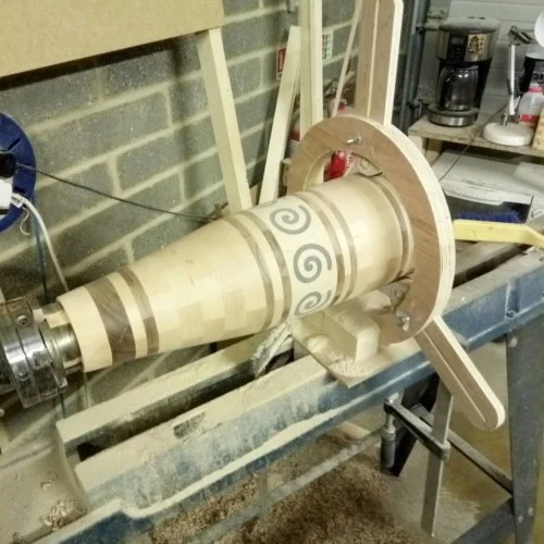 A woodturning lathe with a partially finished wooden spiral piece mounted on it, situated in a workshop with brick wall and various tools in the background.