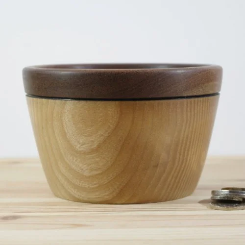 A wooden bowl with a dark brown rim, placed on a light wooden surface beside some coins.