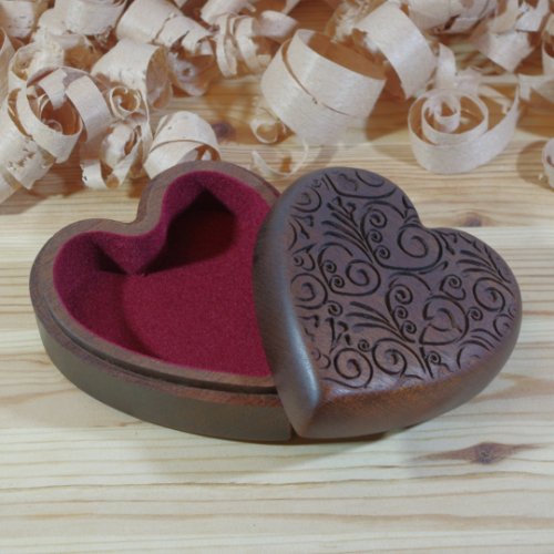 A heart-shaped wooden box with carved intricate patterns on the lid and a red velvet interior, surrounded by wood shavings.