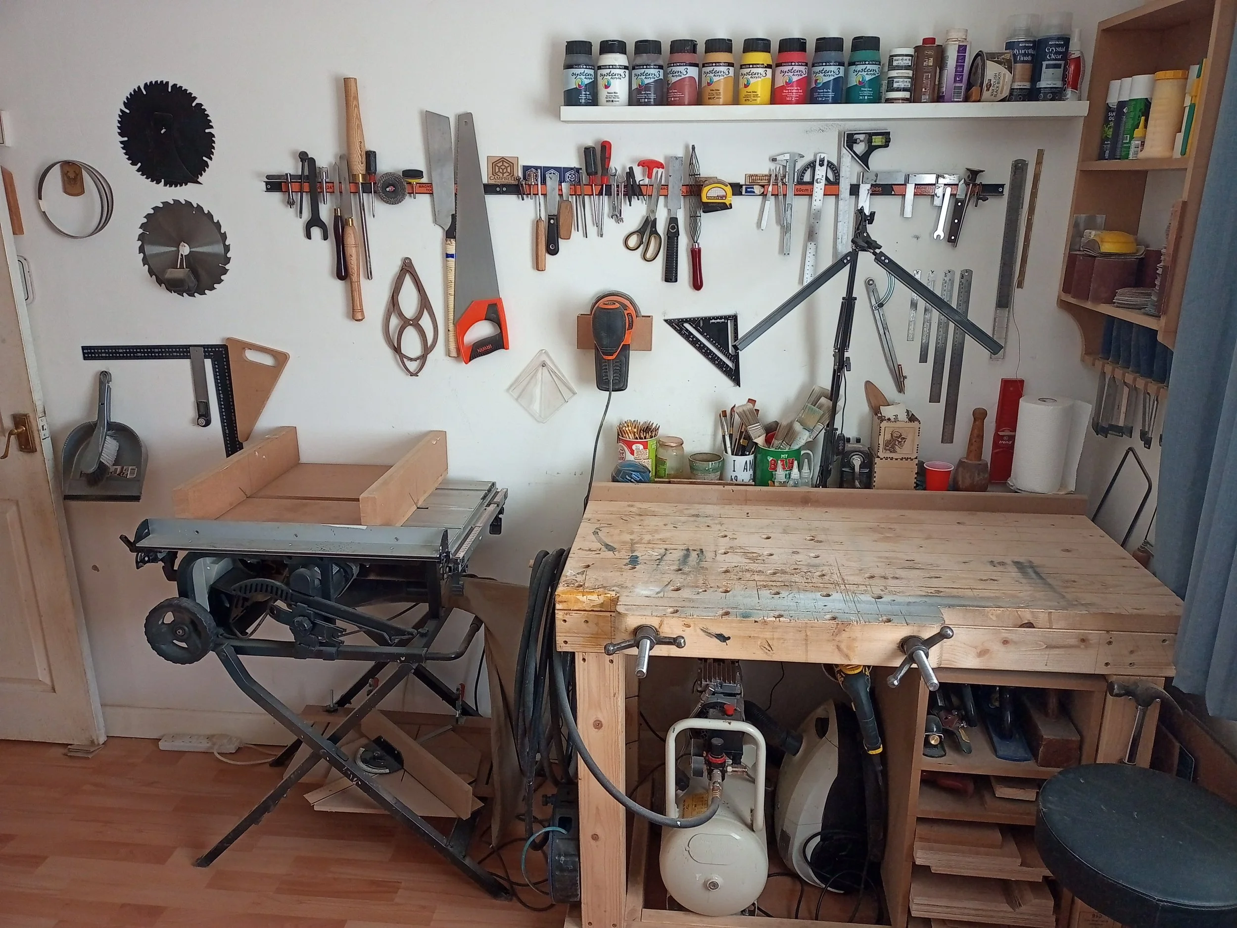 A woodworking workshop with a workbench, various tools hanging on the wall, a table saw, and numerous paint pots on a shelf.