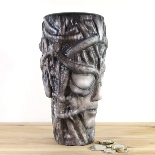 A wooden vase inspired by H.R.Giger resembling a human face with intertwined pipes and features, placed on a wooden surface with some coins in front.